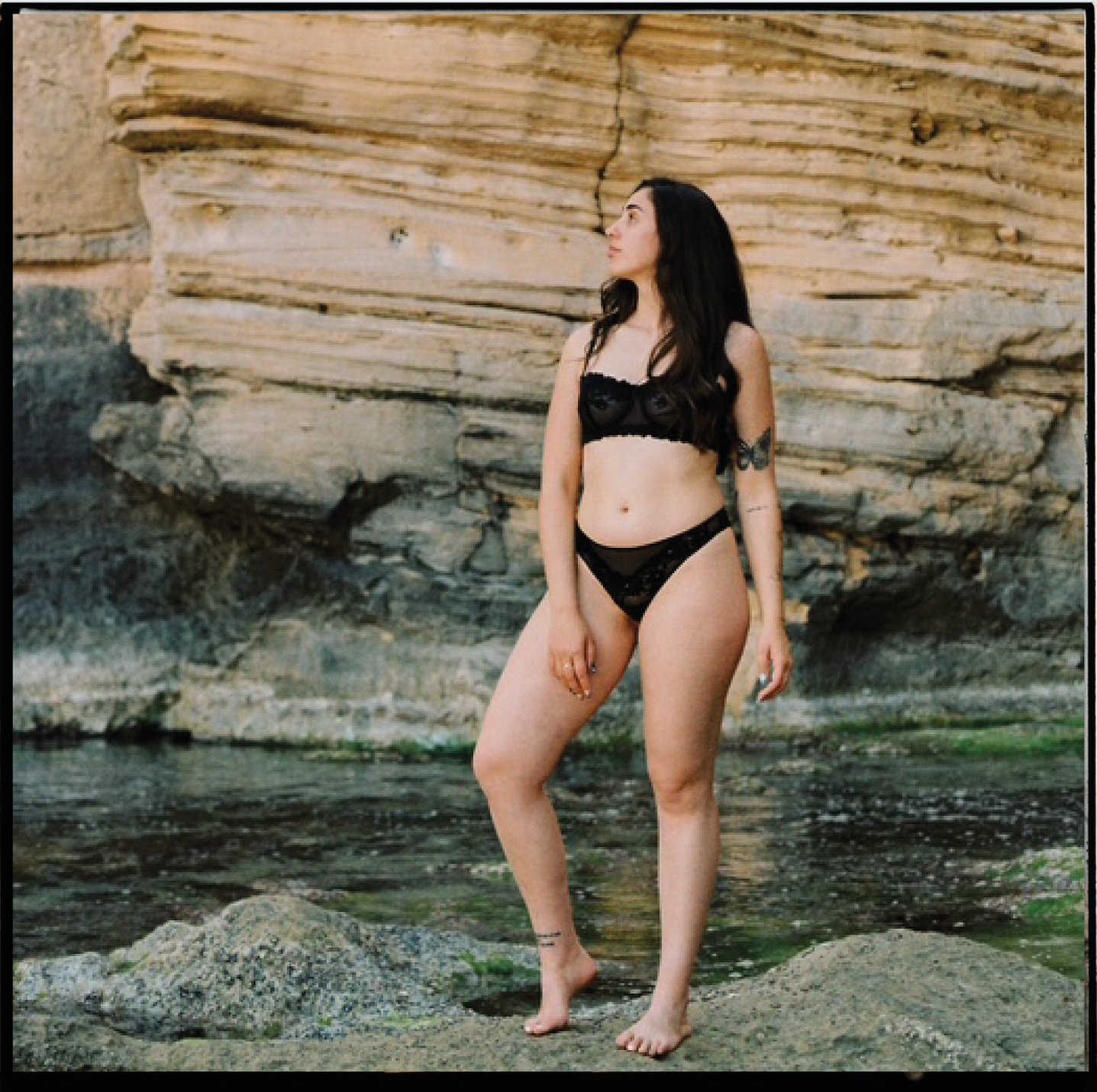Mujer con ropa interior negra de encaje, de pie en rocas junto a agua, con pared de piedra en el fondo.