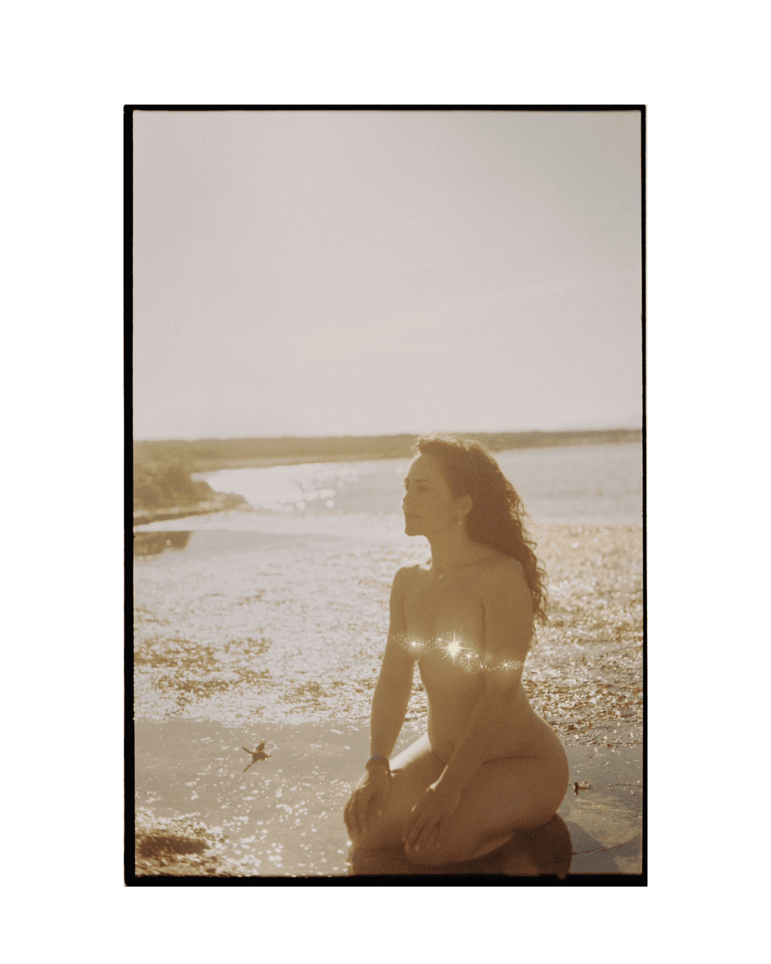 Mujer desnuda con cabello rizado en la playa durante el atardecer, cubierta parcialmente con destellos de luz.