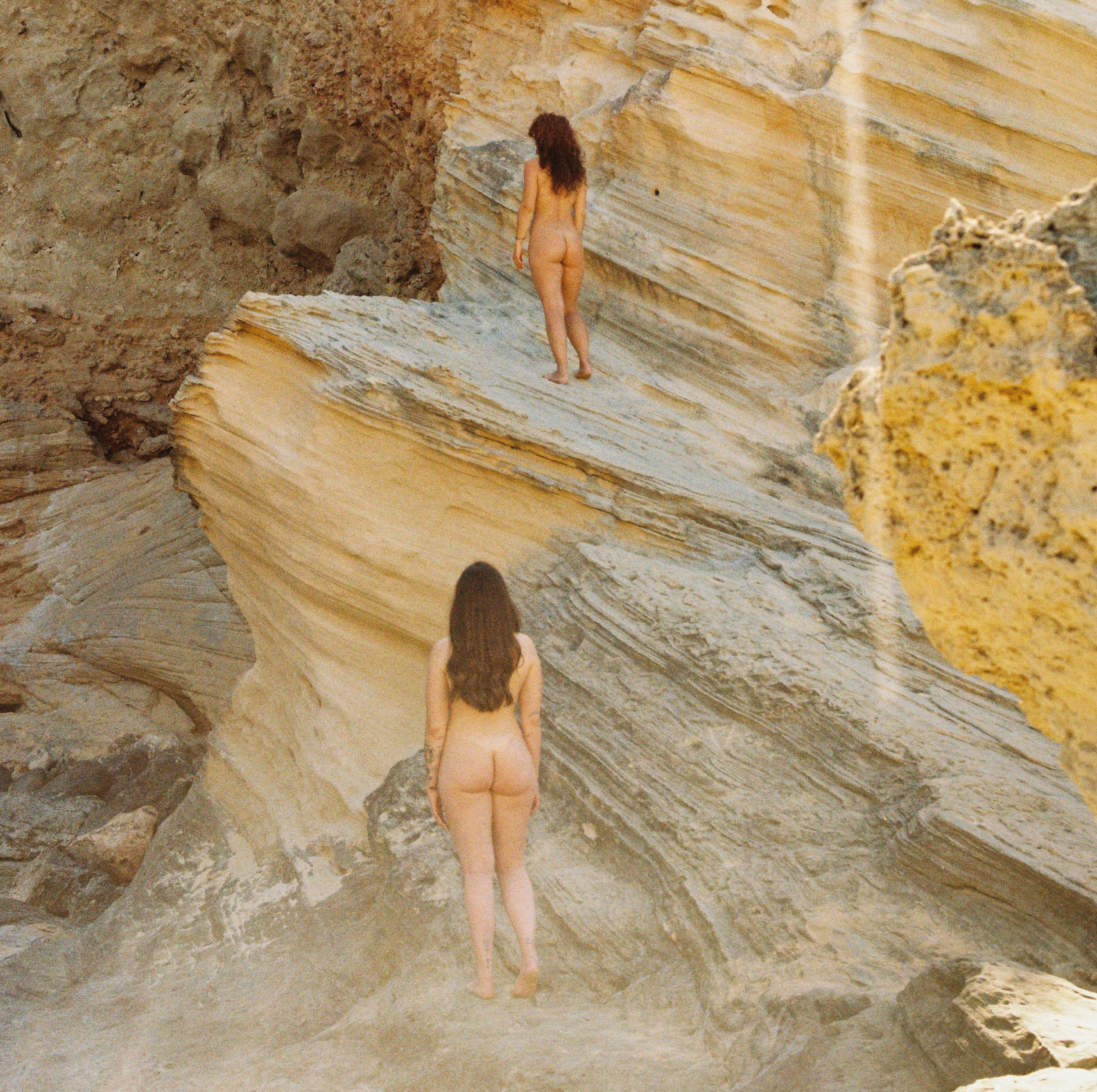 Una mujer desnuda con cabello largo y rizado camina sobre una formación rocosa en un entorno natural con rocas de colores beige y marrón.