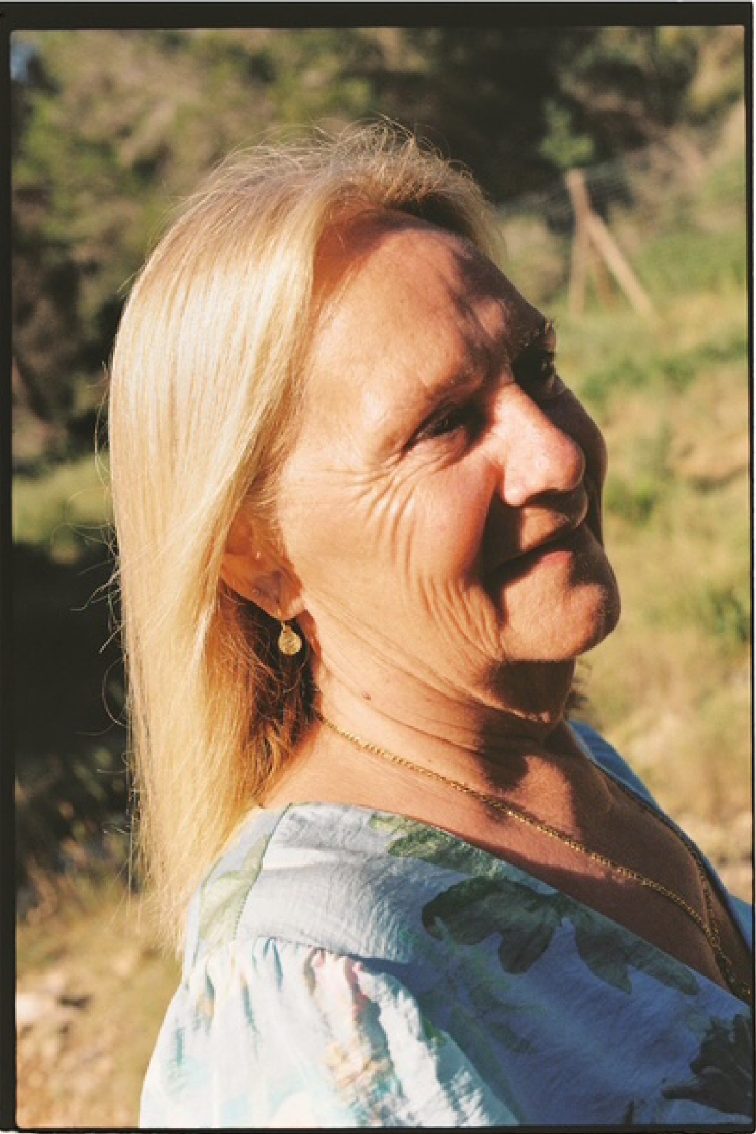Retrato de mujer mayor con cabello rubio y largo, con arrugas y usando pendientes y collar, sonriendo al sol en un entorno natural