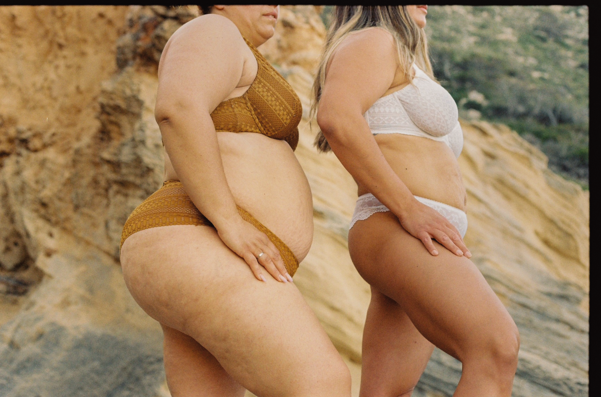 Captura de dos mujeres en traje de baño, de espaldas, en un entorno al aire libre con rocas y vegetación.