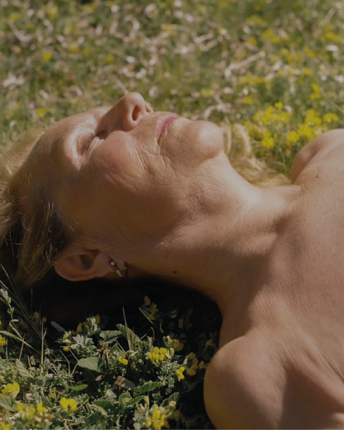 Mujer joven durmiendo en campo con flores y árboles en el fondo con expresión de paz.