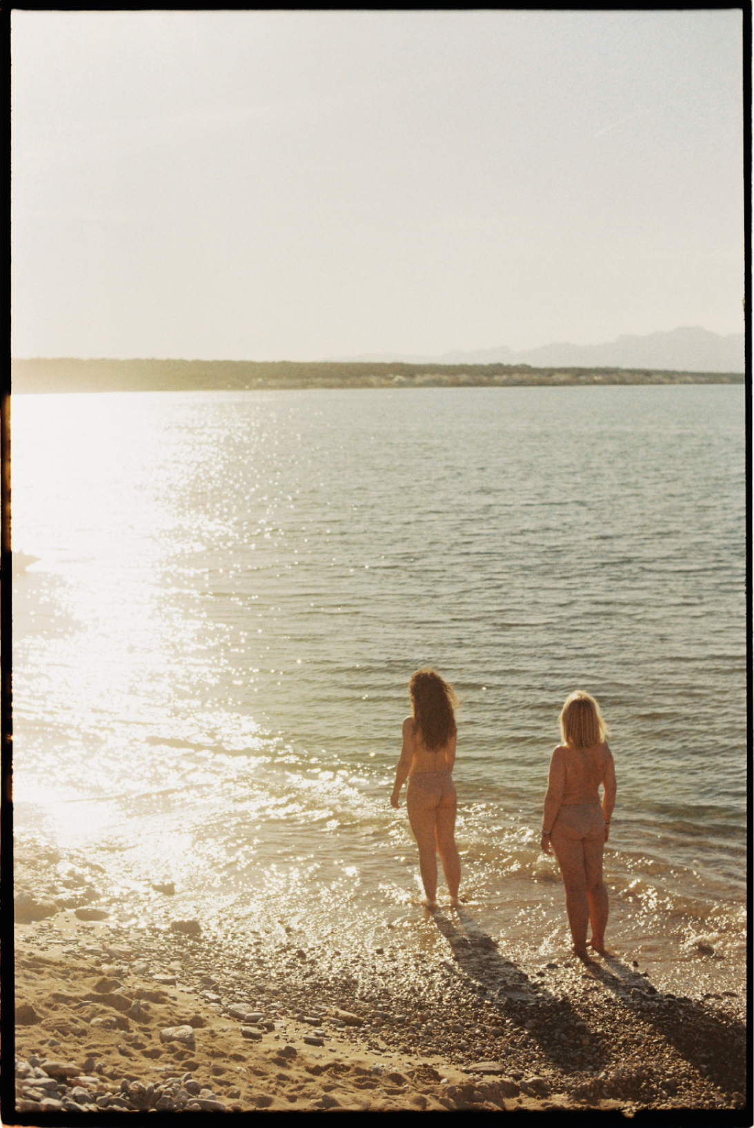 Dos mujeres desnudas caminando por la orilla del mar en una playa durante el atardecer, con el sol reflejándose en el agua y montañas en la distancia.
