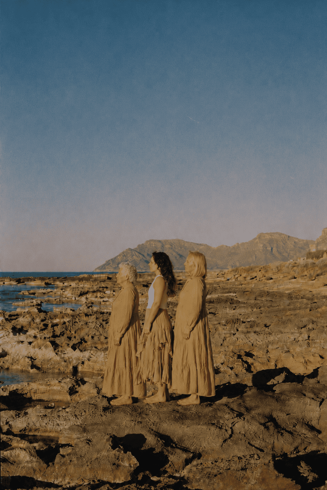 Tres mujeres de diferentes edades de pie en una playa rocosa con montañas al fondo, durante una puesta de sol, vestidas con ropa larga y suelta de color beige.