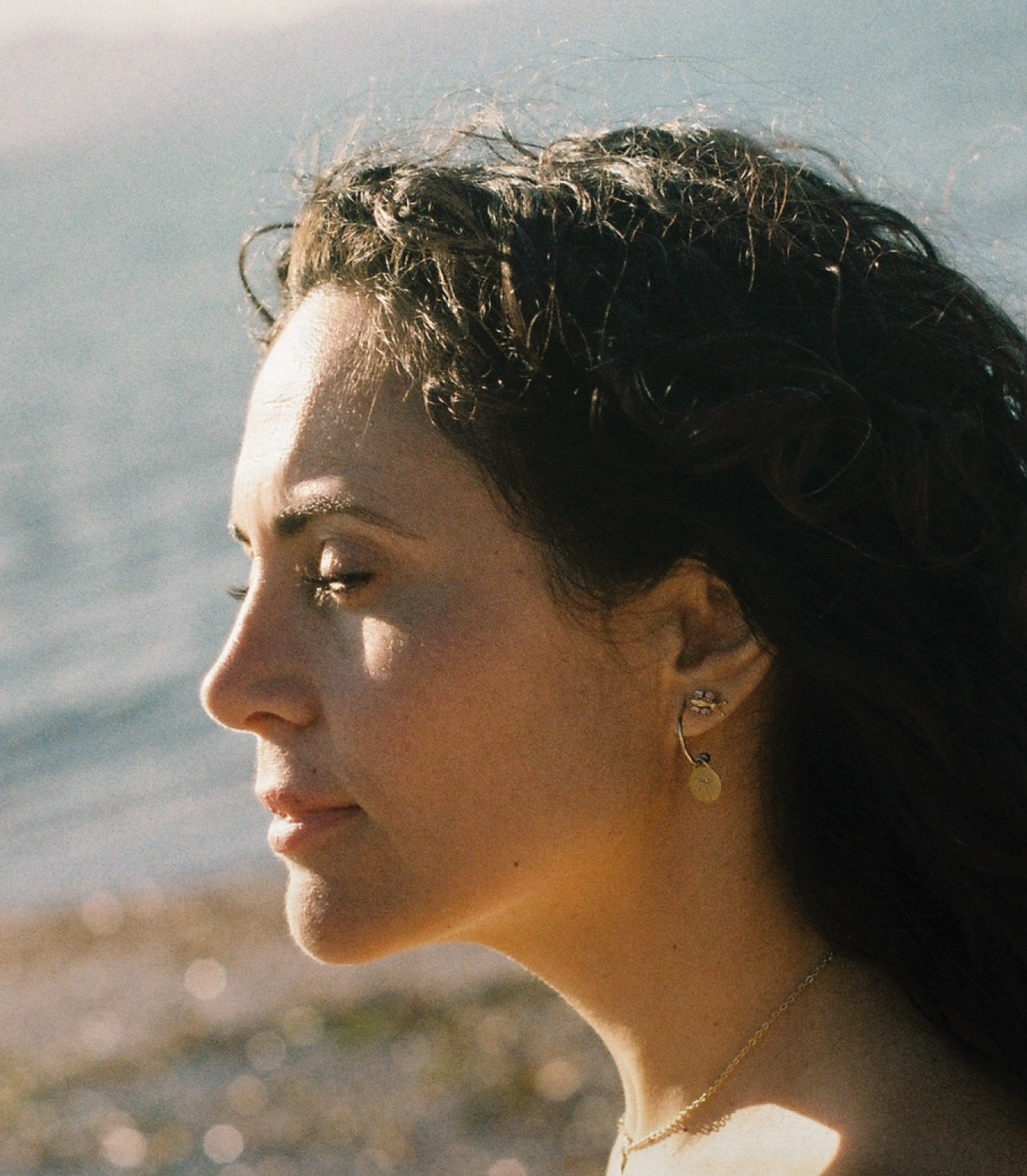 Retrato de perfil de una mujer con cabello oscuro y rizado, en un entorno de playa con el mar al fondo.