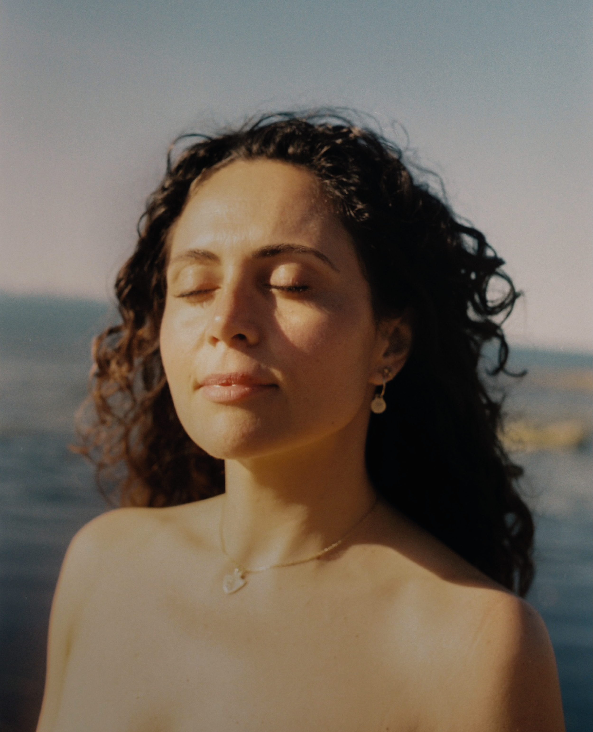 Retrato de una mujer con ojos cerrados, cabello rizado y expresión tranquila, en un entorno exterior con el mar de fondo.