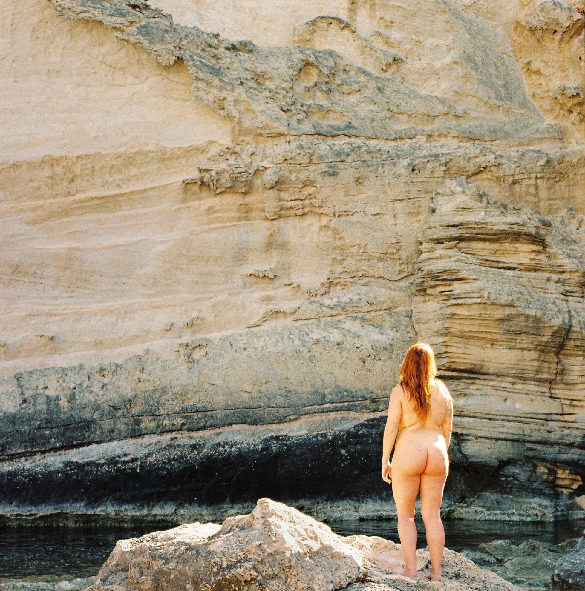 Mujer desnuda con cabello largo en la orilla de un río, mirando un muro de piedra.