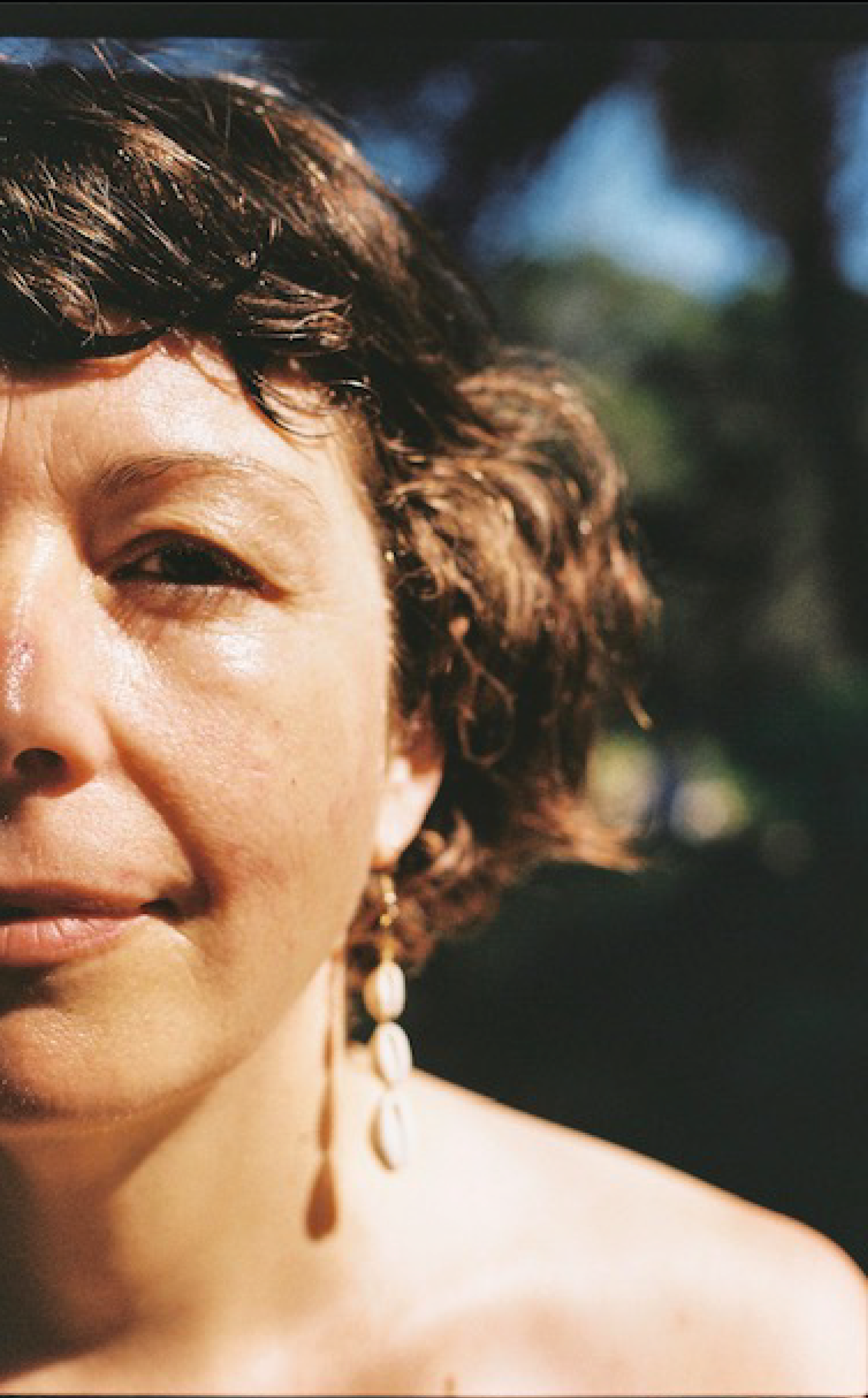 Primer plano del rostro y el cabello de una mujer de piel clara, con pendientes colgantes y cabello rizado y castaño oscuro. Fondo desenfocado con cielo y árboles.
