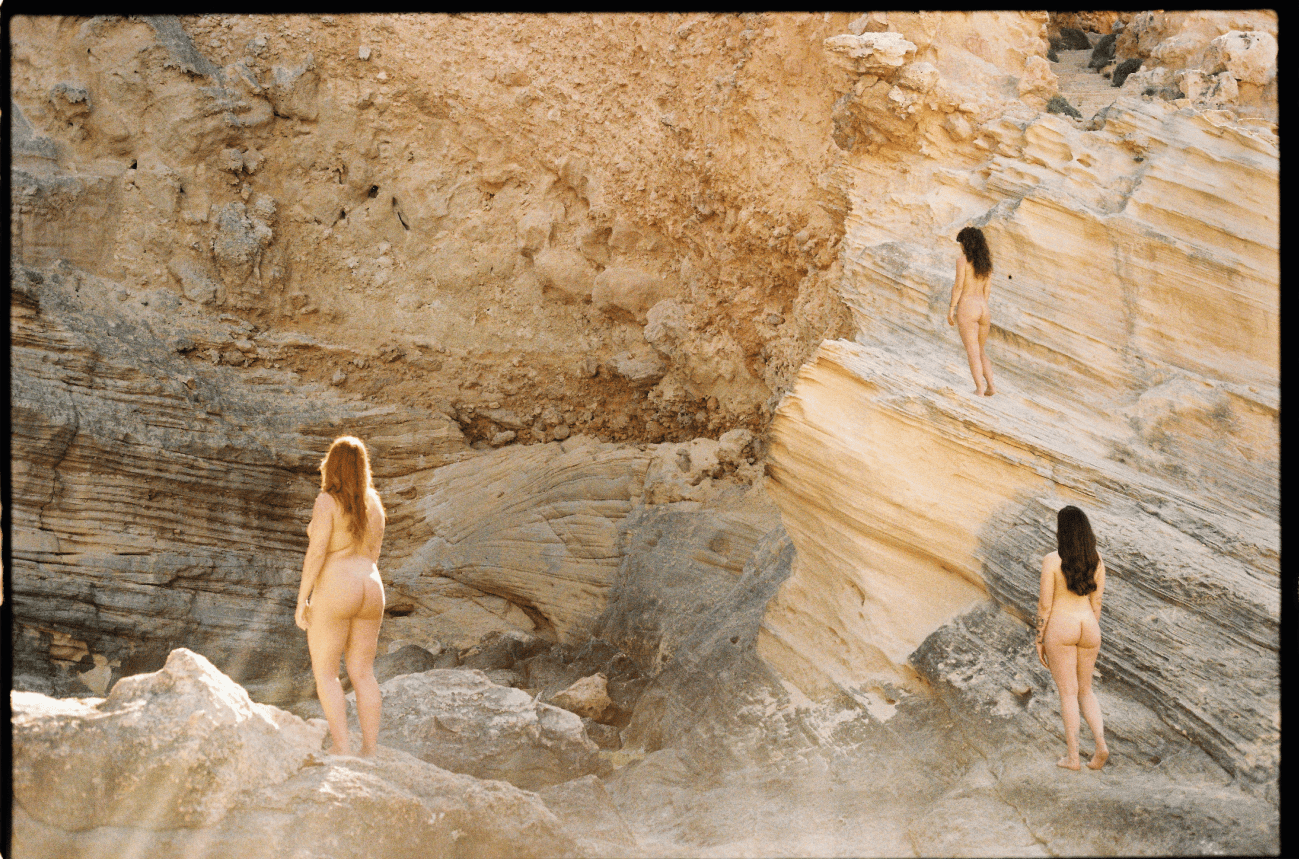 Tres mujeres desnudas caminando sobre formaciones rocosas en un entorno árido y rocoso.