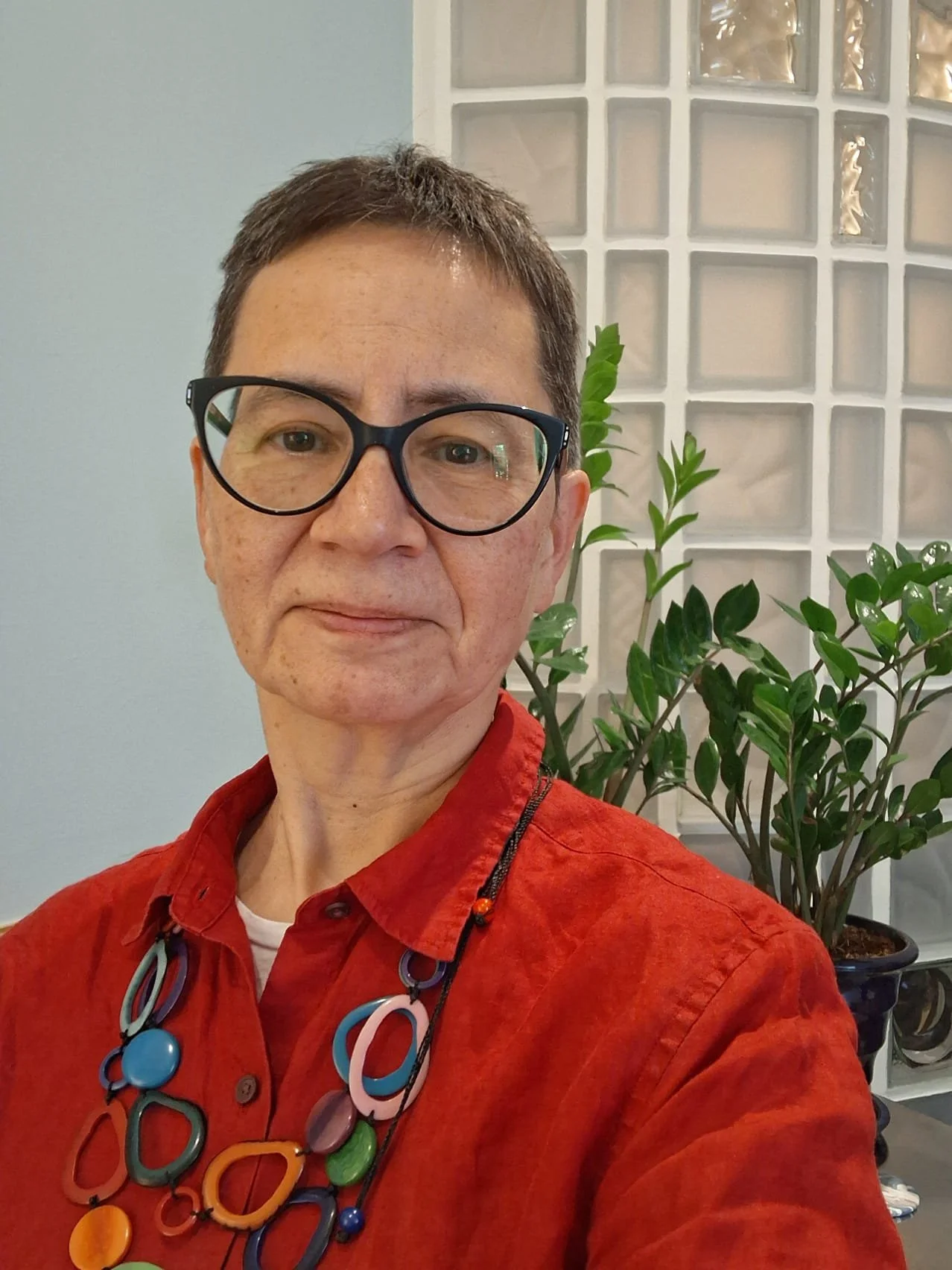 A woman with short brown hair and glasses wearing a red shirt and colorful necklace. She is indoors, standing in front of a glass brick wall and a potted green plant.