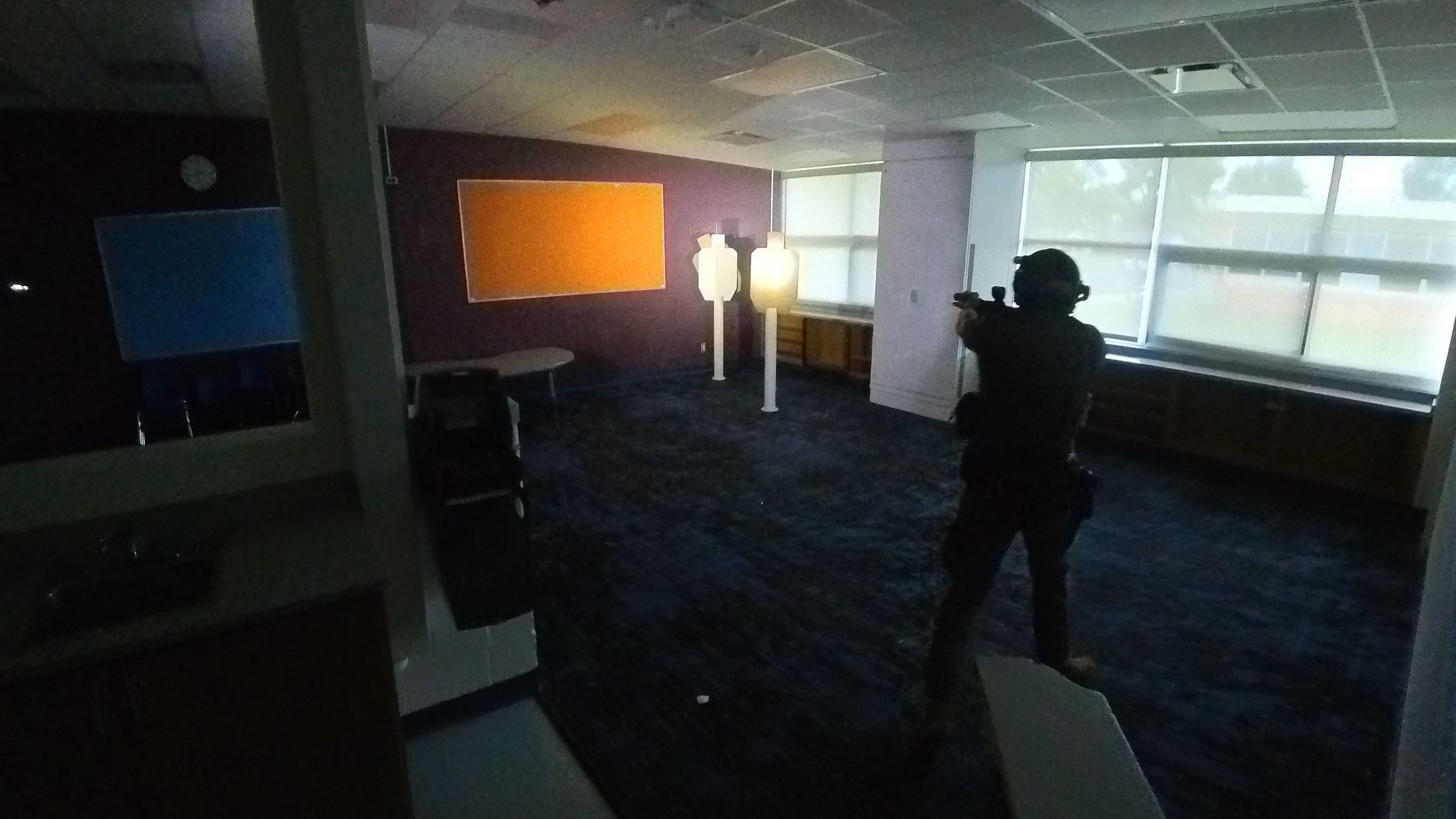 A person wearing a helmet and dark clothing is aiming a rifle with a scope inside a dimly lit room with large windows. The room has a purple wall with an orange rectangular painting, two white lamps, a table, and some chairs.