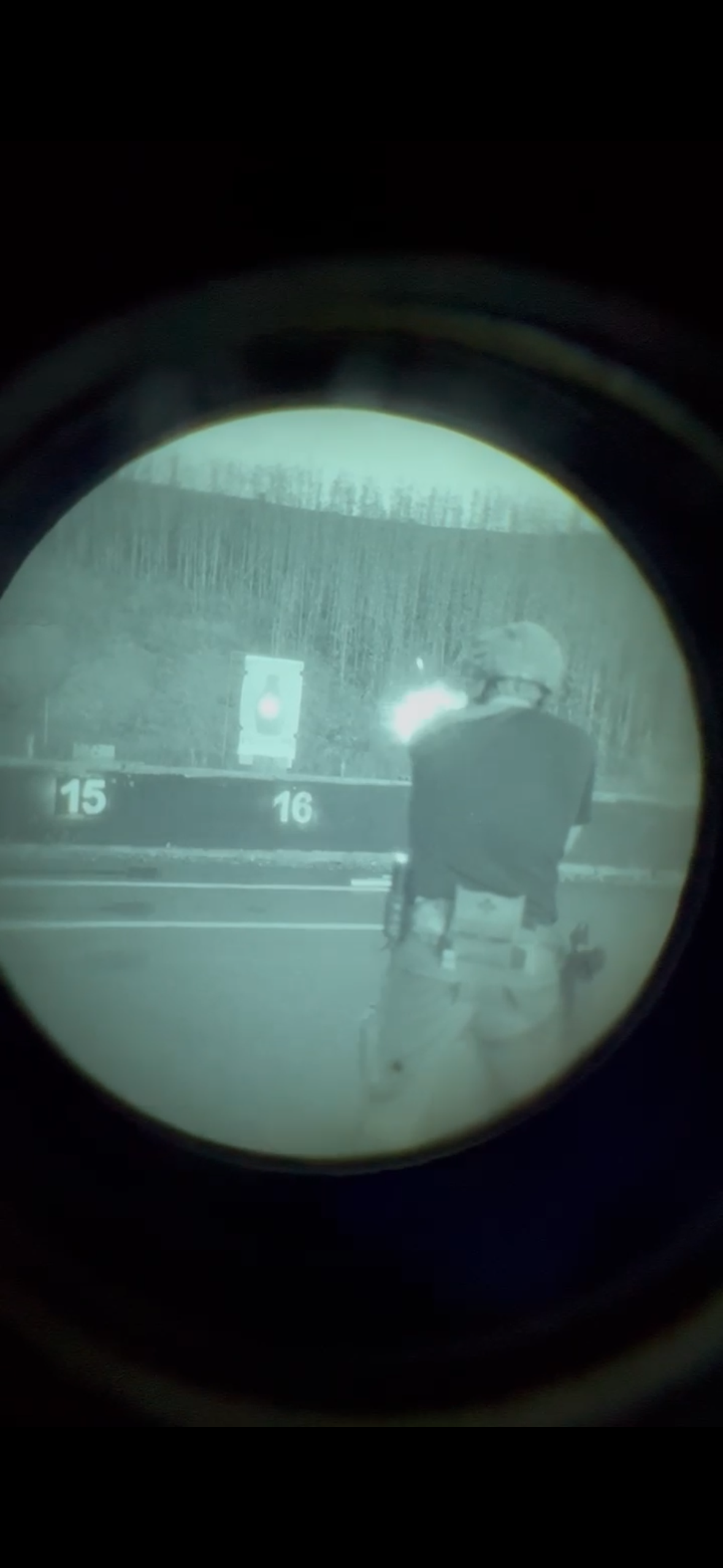 Person aiming a firearm at a shooting range, viewed through a scope, with target boards numbered 15 and 16 in the background.