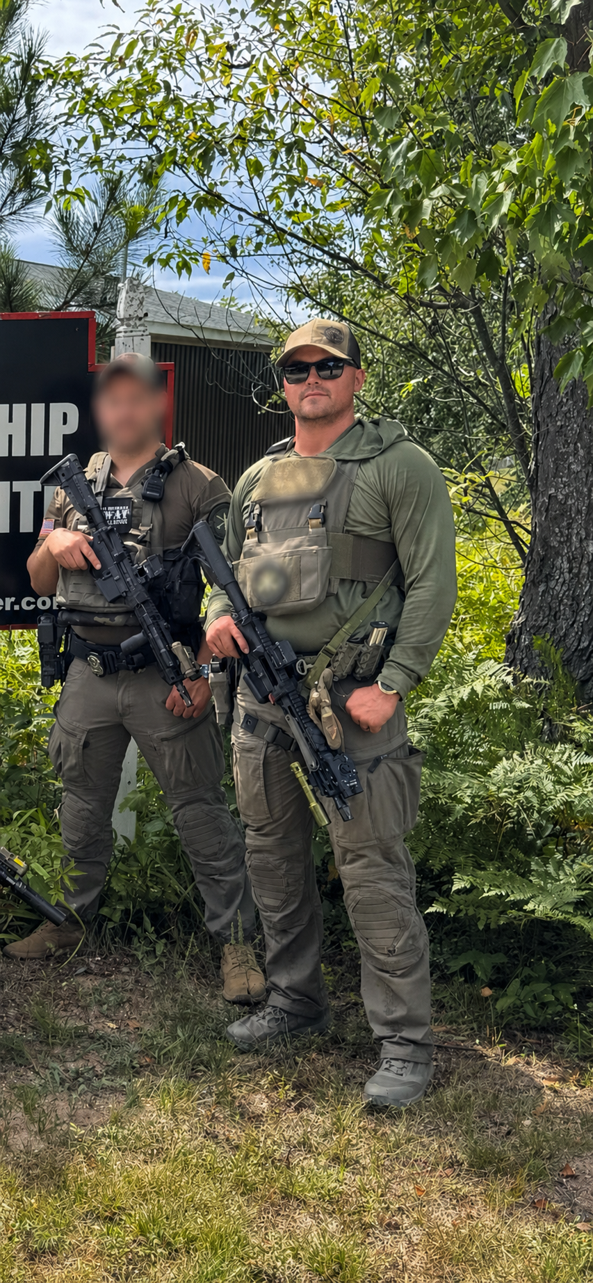 Two armed men in tactical gear standing outdoors among trees and bushes, with one man wearing sunglasses and a cap.
