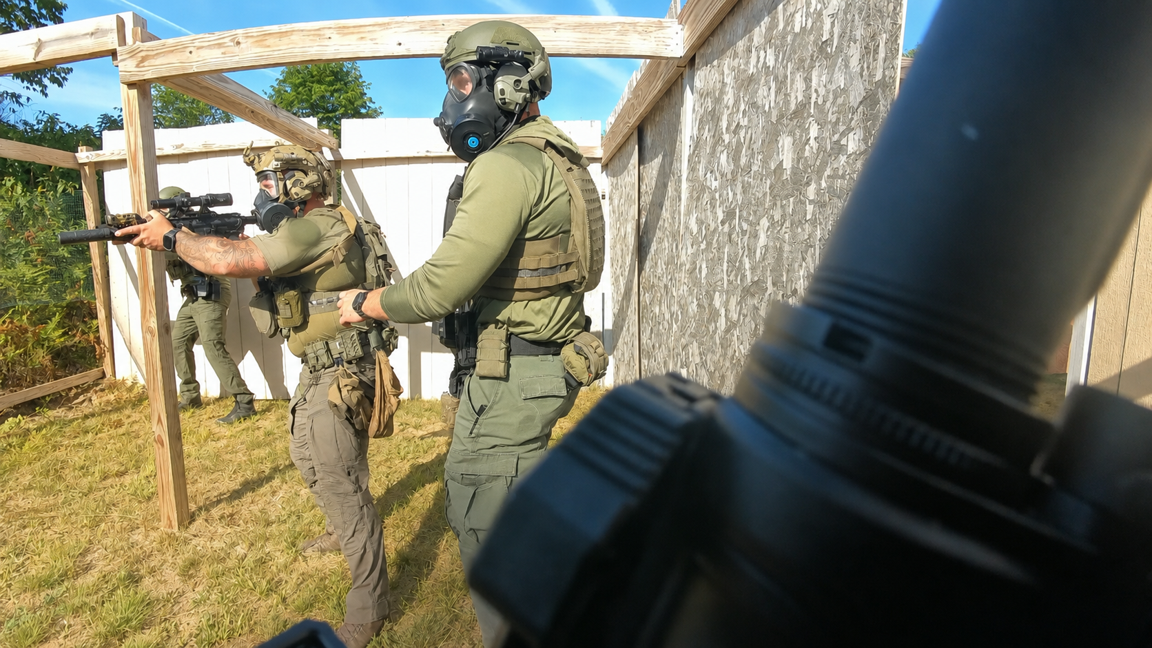 Three armed military personnel in tactical gear holding rifles, with one person aiming their weapon in an outdoor training or simulation area with a wooden and plywood structure, fences, and trees in the background.