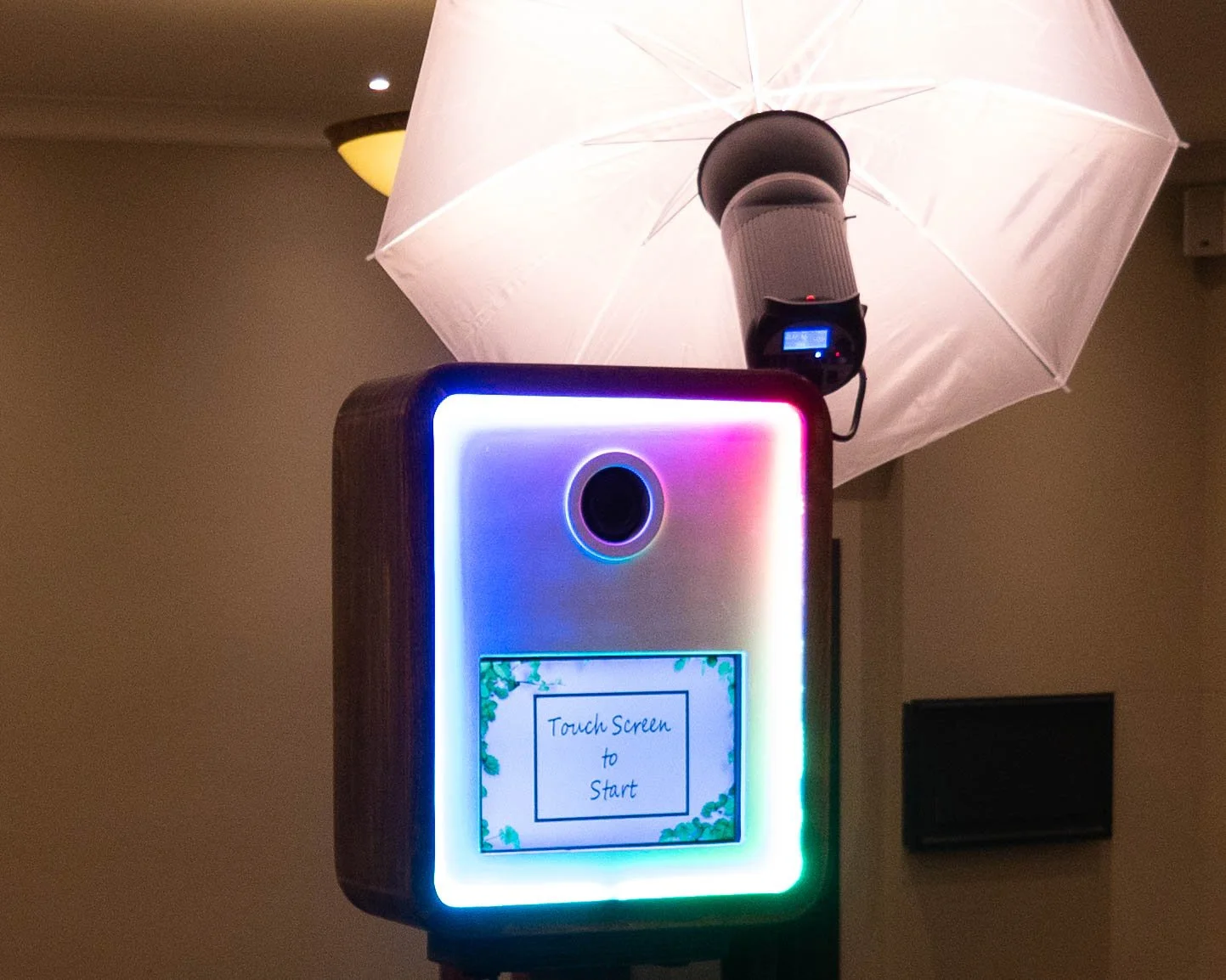 A photo booth with rainbow LED lights around the border, a digital screen with the message "Touch Screen to Start," a camera lens above the screen, and an umbrella-style light diffuser with a camera attached, in a dimly lit room.