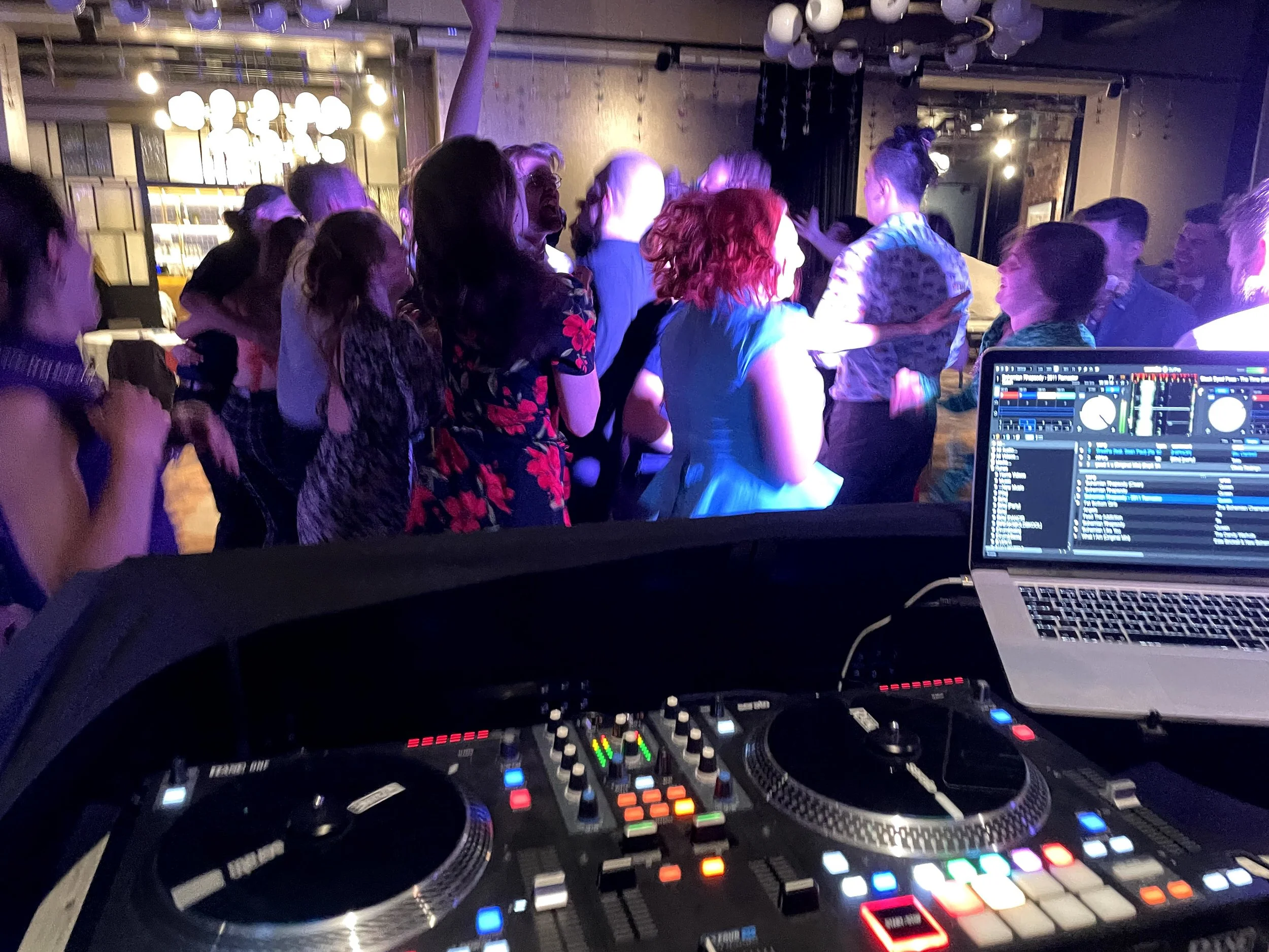 A DJ booth inside a nightclub with a DJ's laptop, mixer, and controller in the foreground. Behind, a group of people are dancing and socializing with colorful lighting and decor.