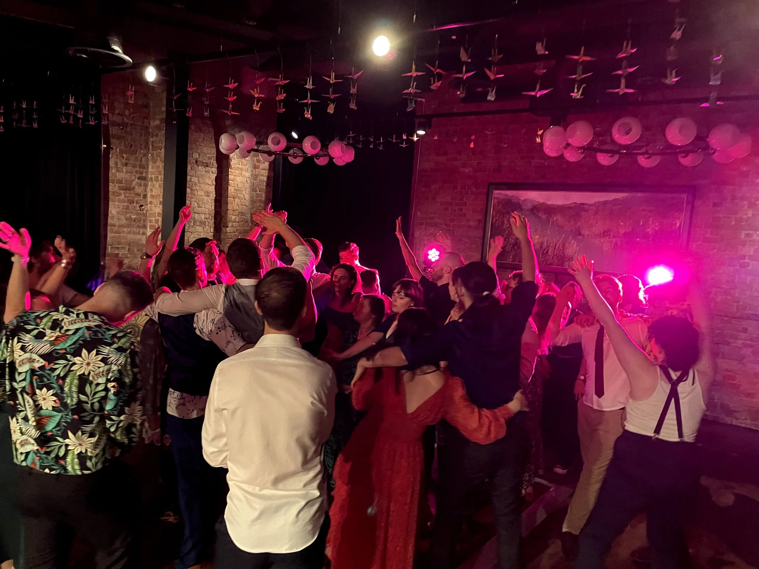 People dancing at a lively indoor party with colorful lighting, suspenders hanging from the ceiling, exposed brick walls, and a vibrant atmosphere.