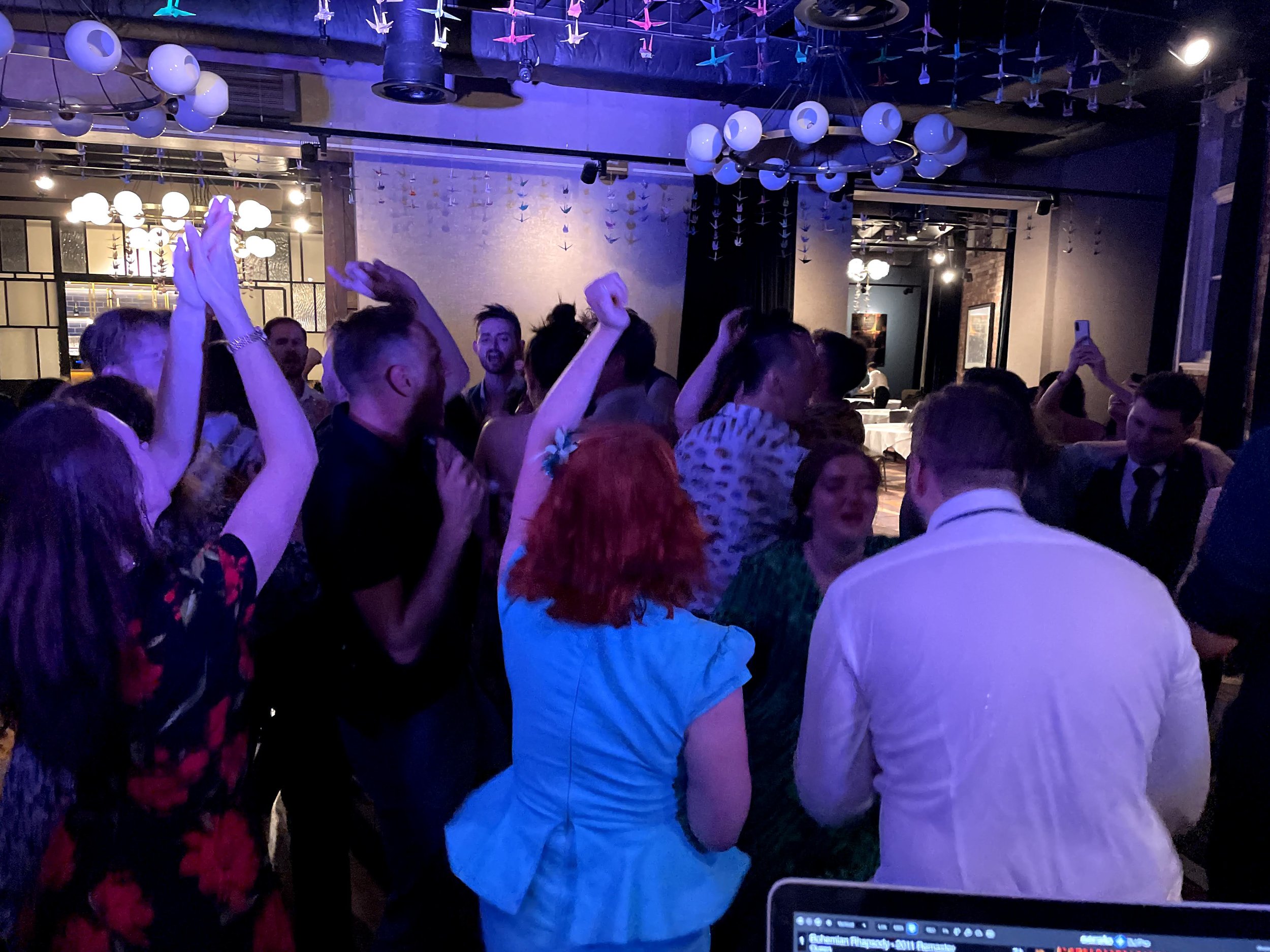 People dancing and celebrating in a dimly lit venue with hanging paper cranes and decorative lights.