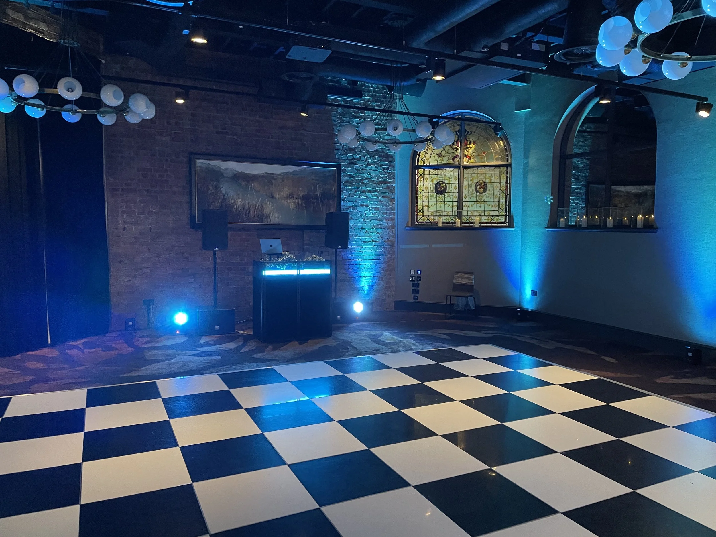 An indoor event space with black and white checkered flooring, exposed brick walls, and stained glass windows. It features modern lighting fixtures with white globes and blue uplighting on the walls, along with DJ equipment set up for entertainment.