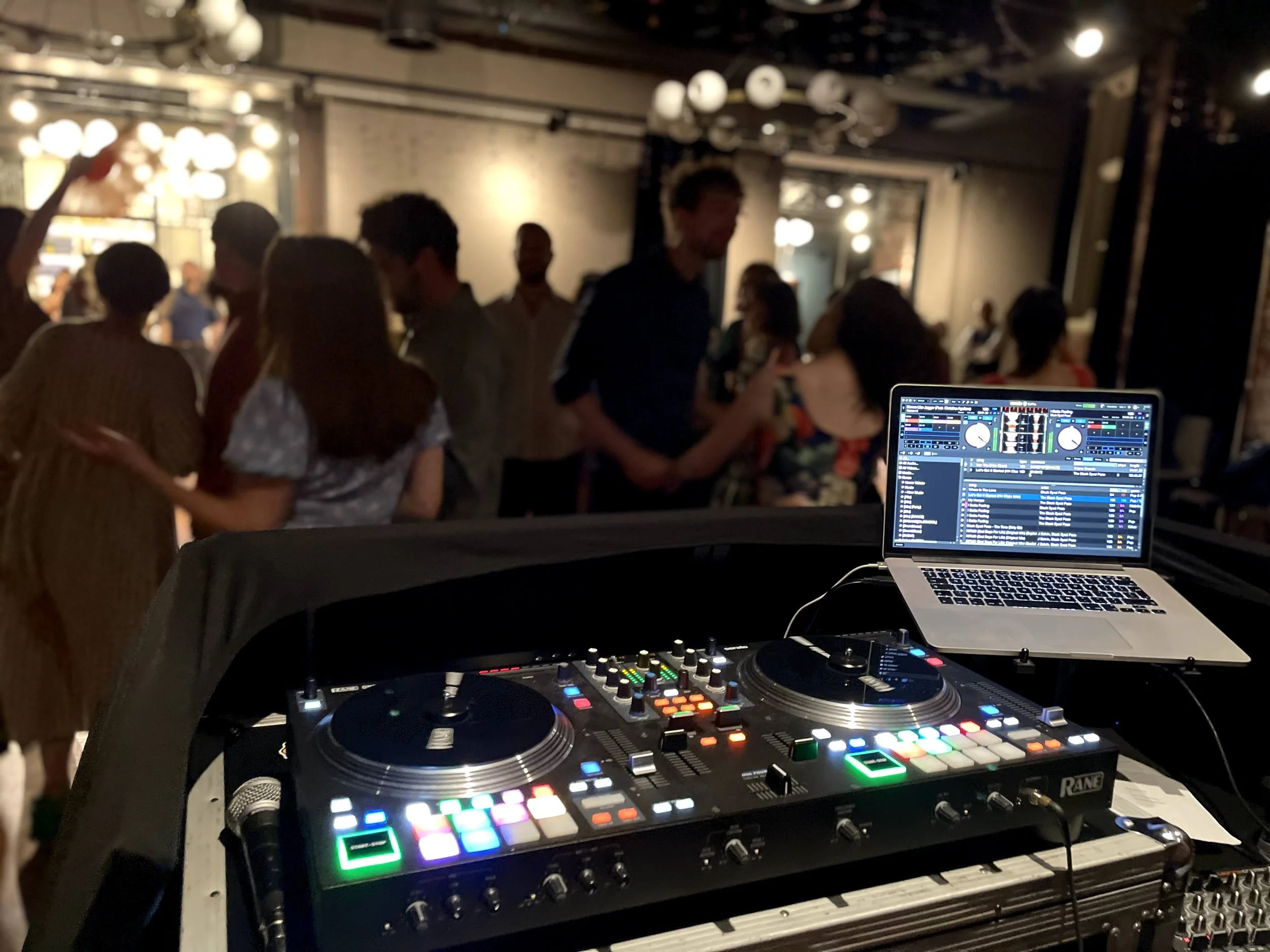 A DJ's mixing console with colorful lights in the foreground, with a crowd of people dancing and socializing in a dimly lit indoor venue, illuminated by hanging globe lights.