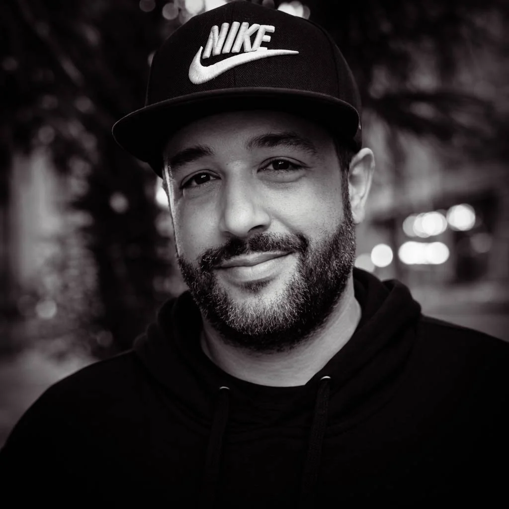 A man with a beard wearing a black Nike cap and black hoodie, smiling at the camera, with a blurred outdoor background.