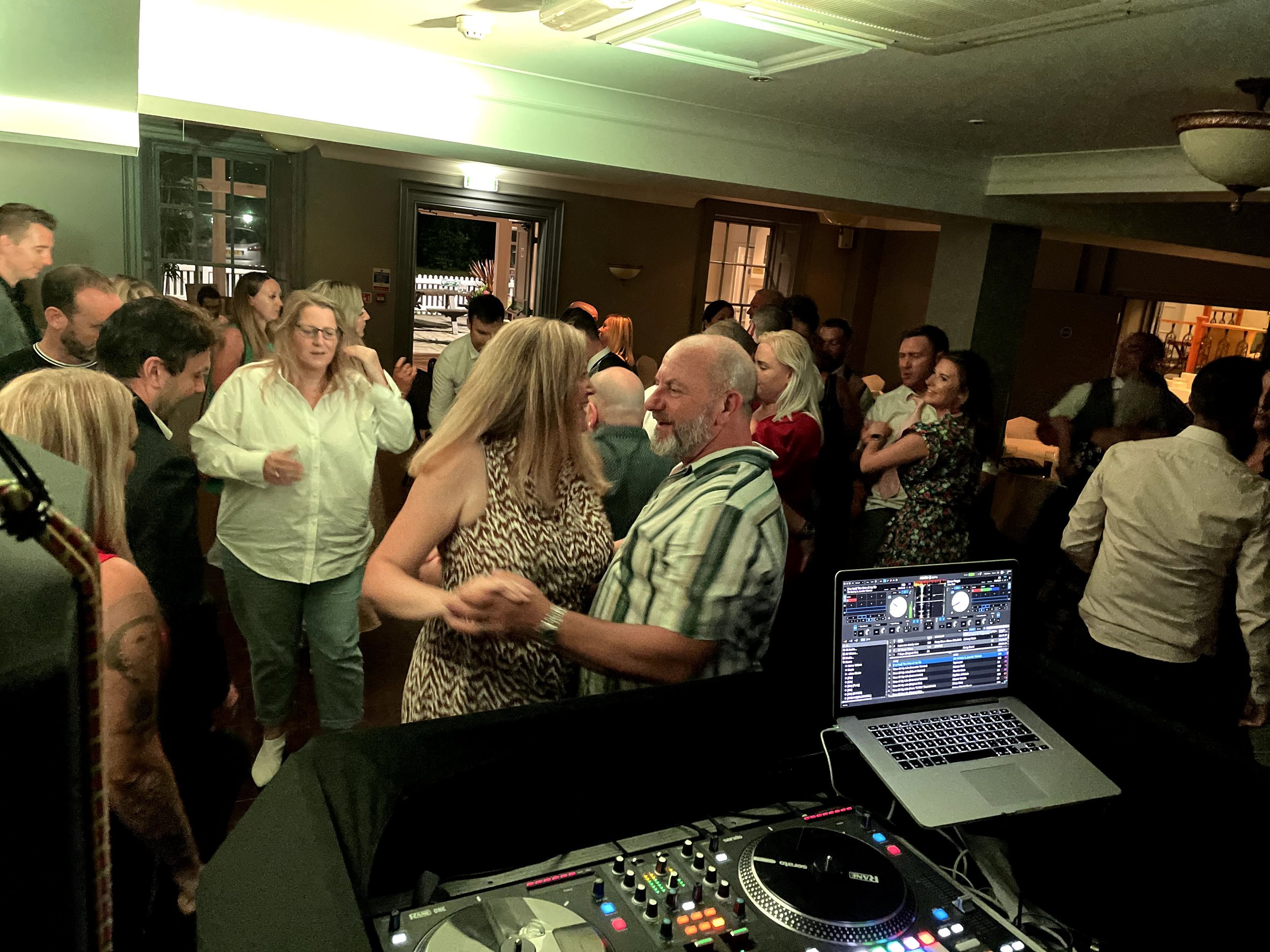 A DJ setup with a laptop and mixing equipment in front of a crowd of people dancing at a party or event in a dimly lit indoor venue.