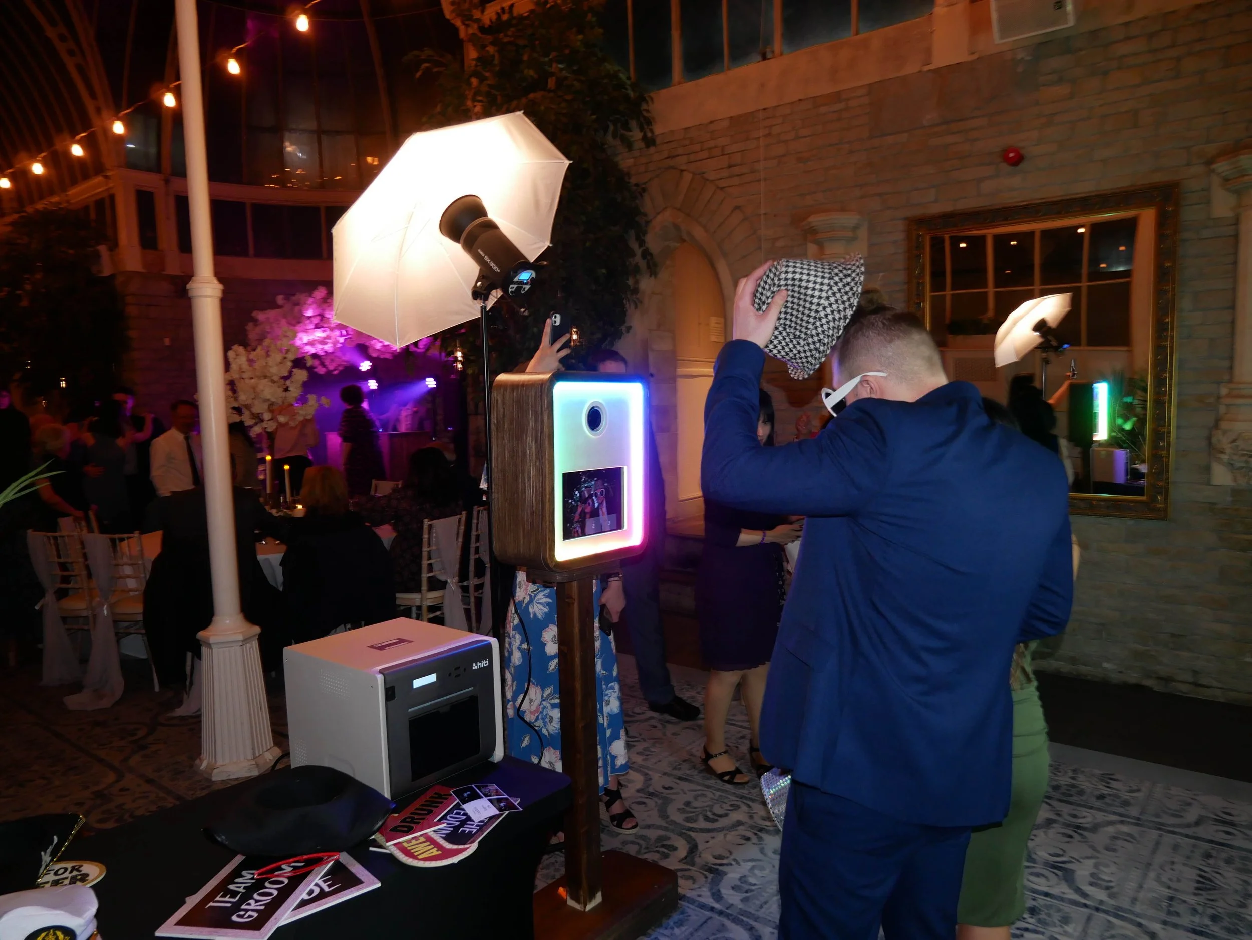 People gathering at an indoor event with photo booth and fun props, including signs and accessories, under warm lighting and decorated with plants and string lights.