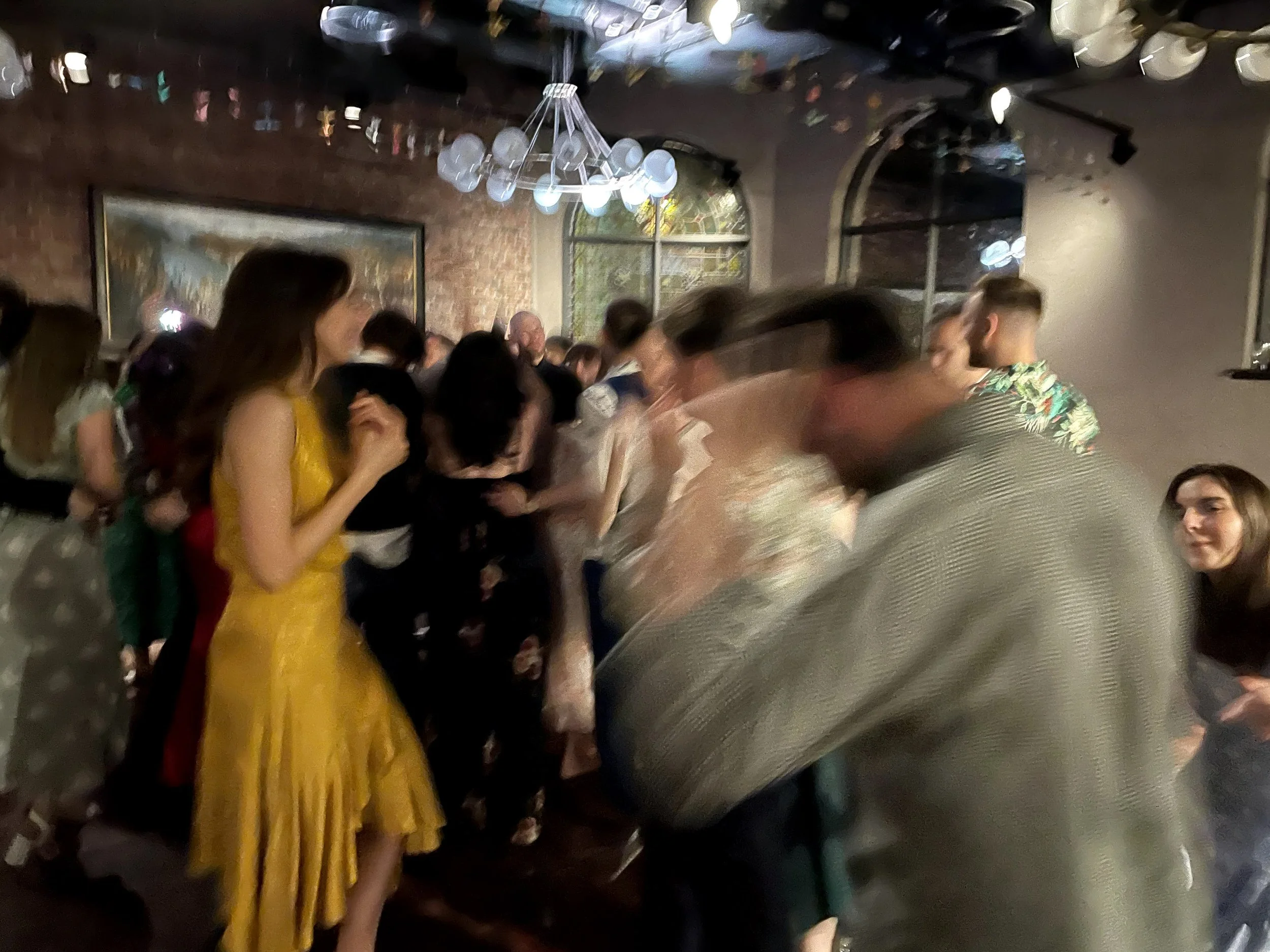A group of people at a social gathering or party, with a woman in a yellow dress in the foreground and others mingling in the background, inside a dimly lit venue with brick walls and large windows.