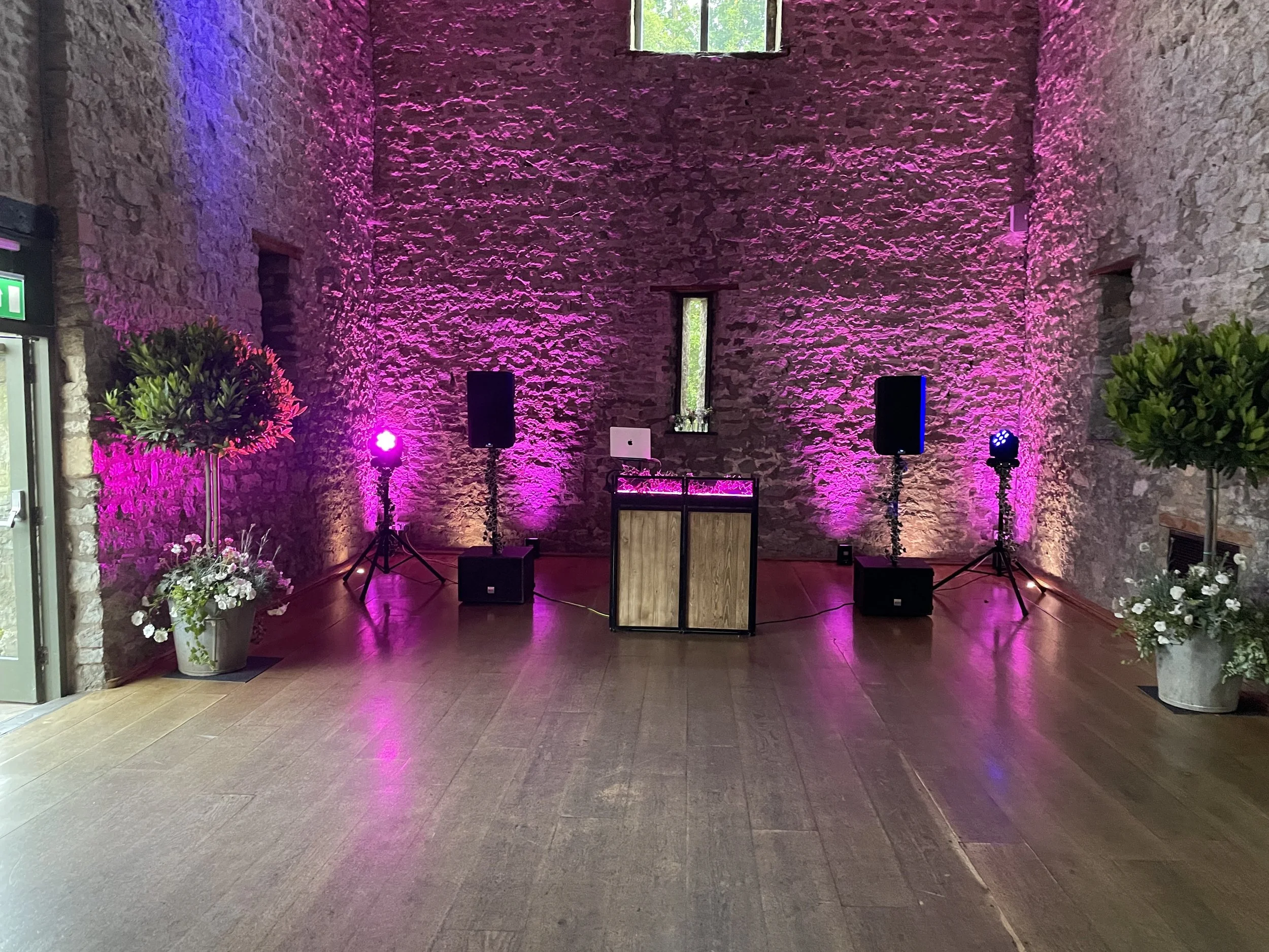 An indoor event space with stone walls illuminated by pink and purple stage lighting. There are two large speakers on stands, a DJ booth with equipment, and decorative plants and flowers on either side of the space.