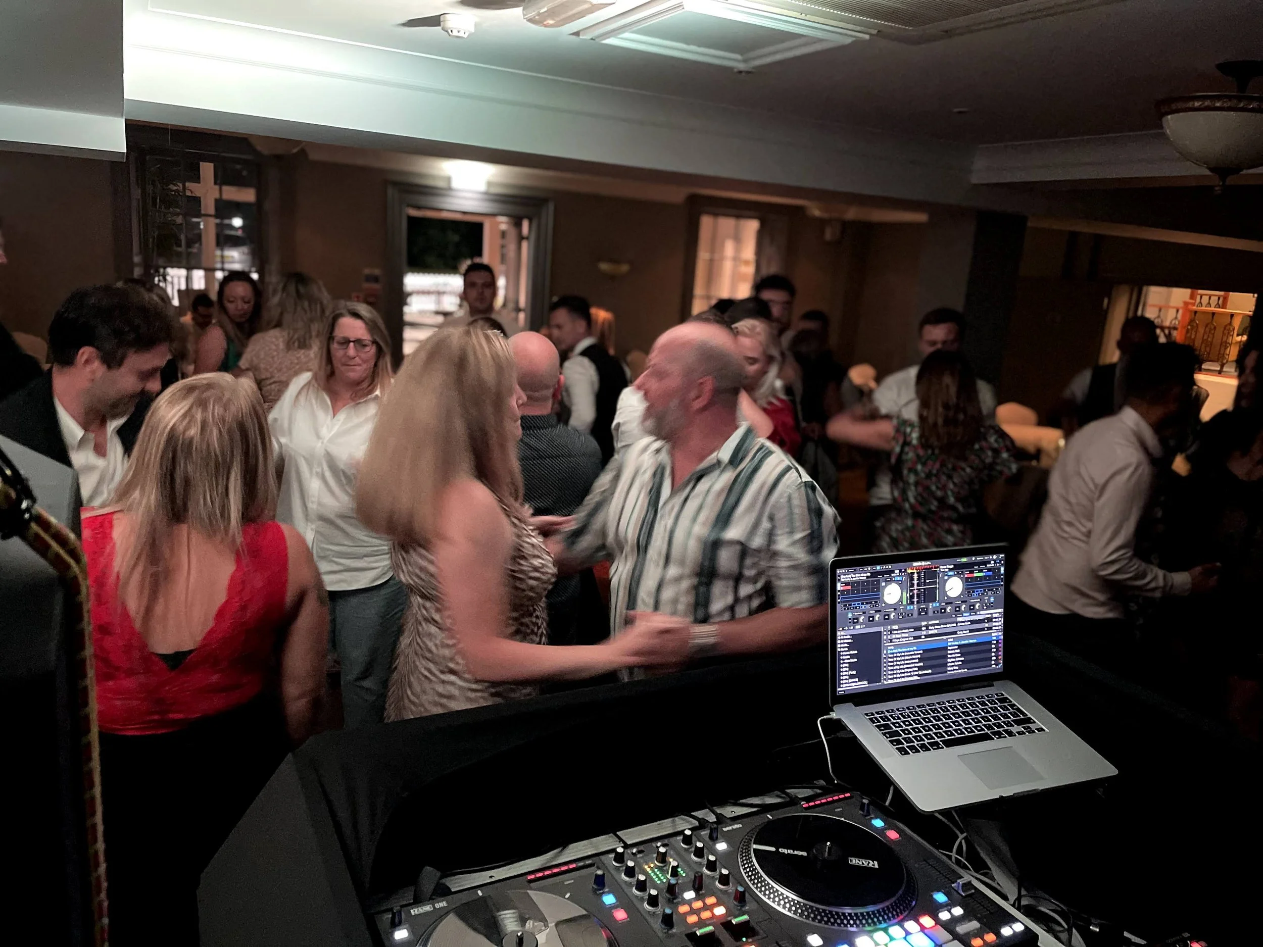 Crowd of people at a social gathering or event, with DJ equipment and a laptop set up in the foreground, creating an energetic atmosphere.