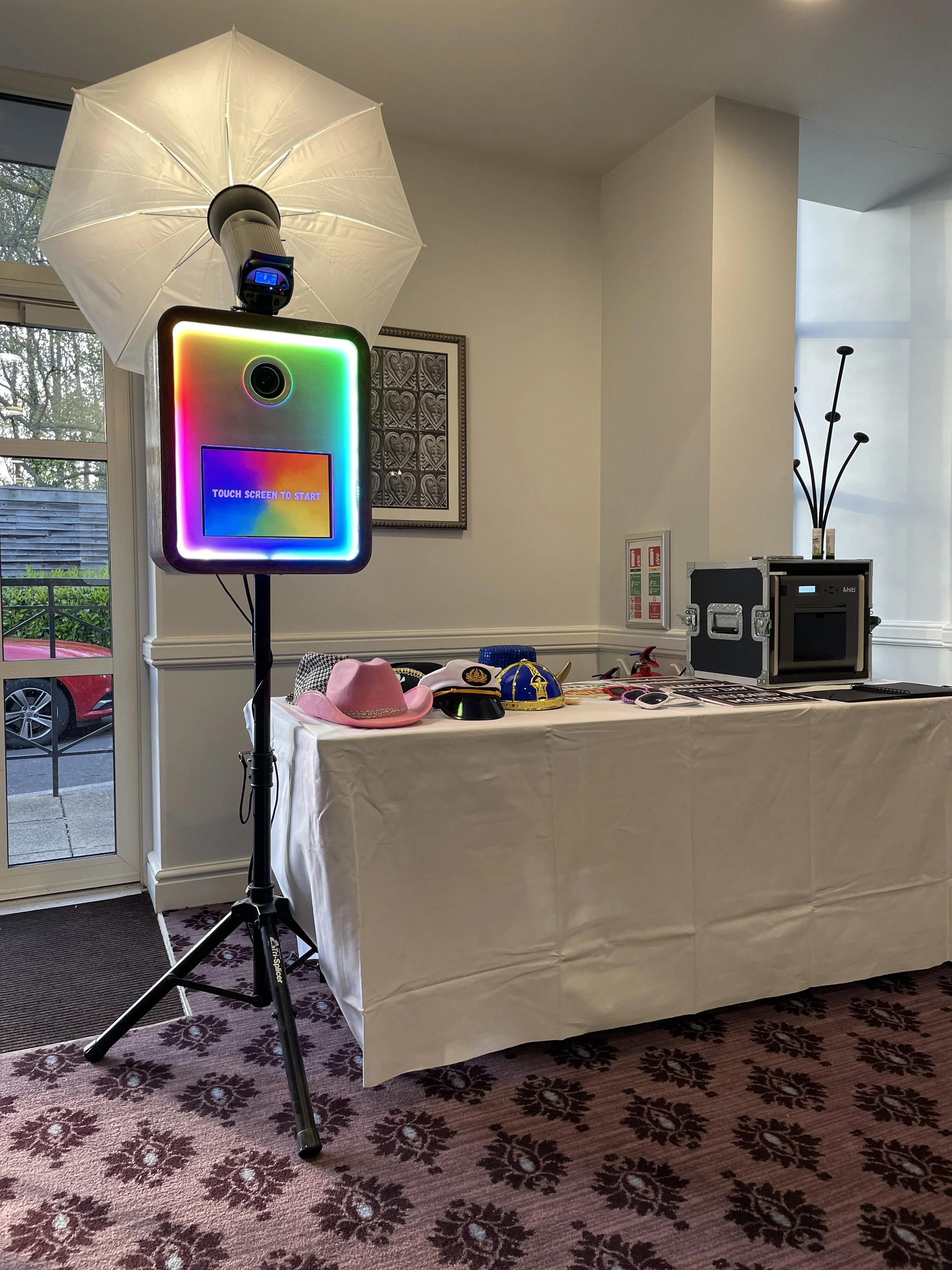 A photo of a room with a table covered with a white cloth, displaying hats and sunglasses. There is a photo booth setup with a colorful illuminated screen and a large umbrella-shaped light. A window and a door lead outside, and there is a decorative 