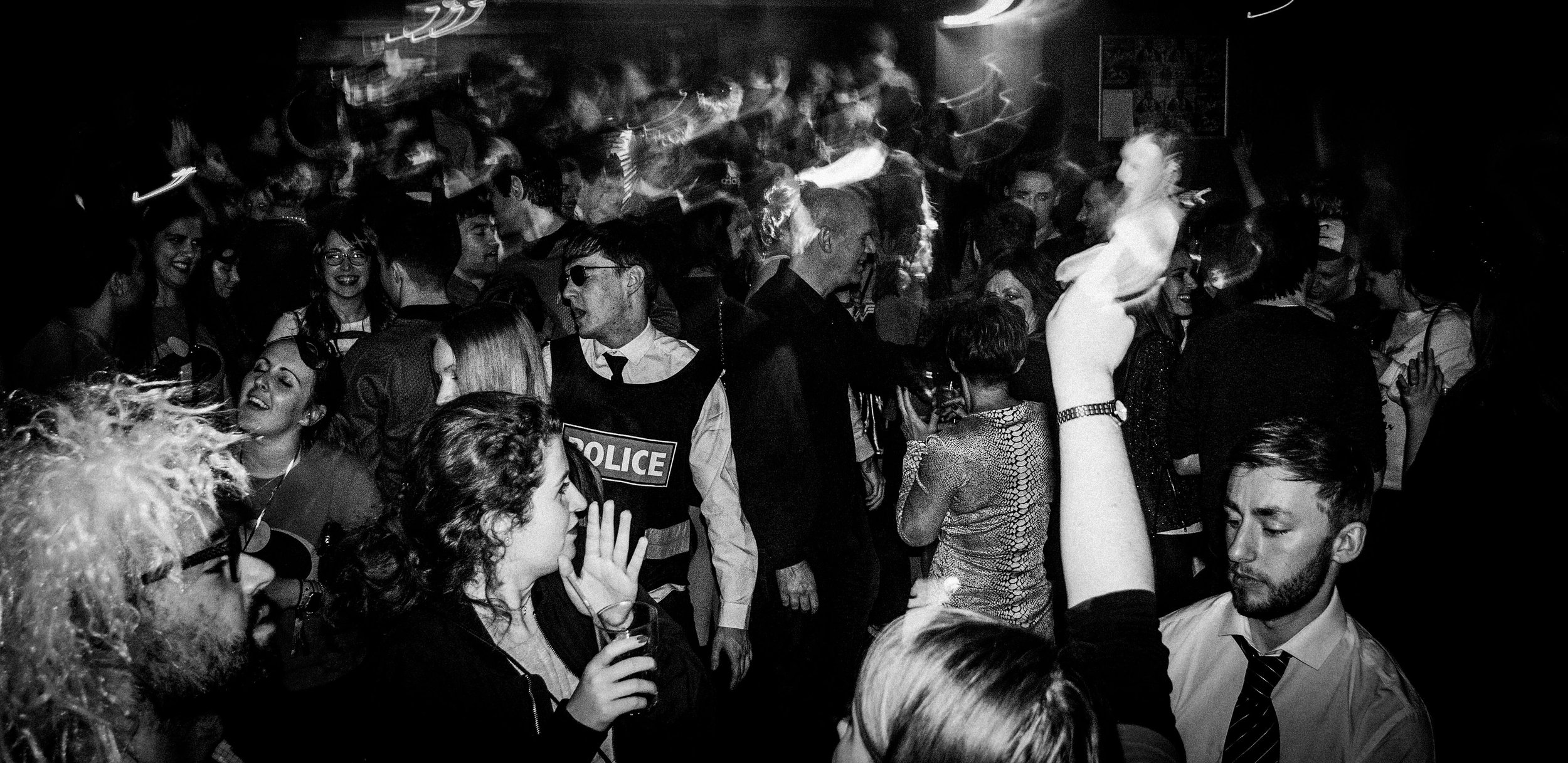 Black and white photo of a crowded dance party with diverse people dancing, socializing, and wearing party attire, including police costumes on some.