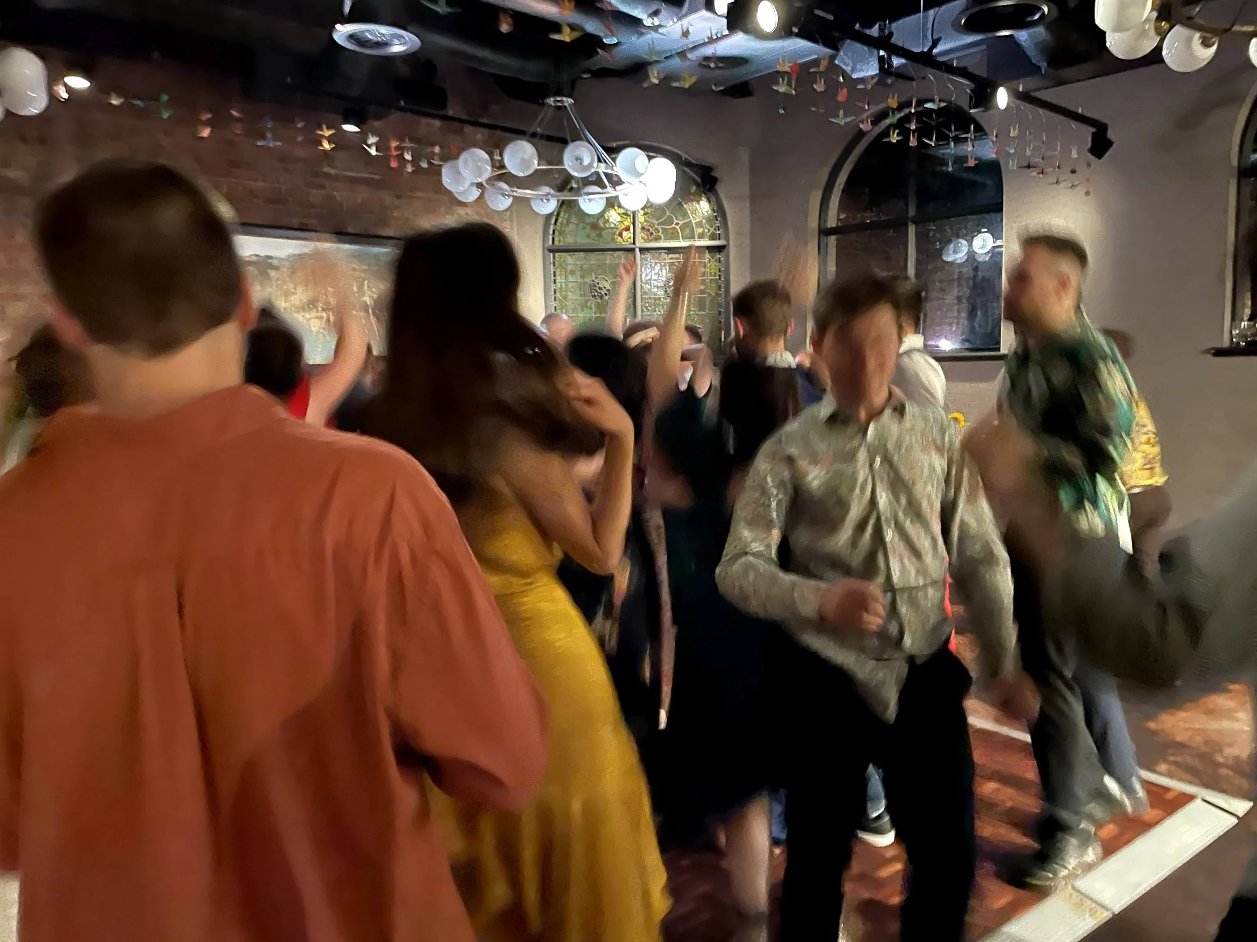 A lively party scene with people dancing in a dimly lit room, decorated with colorful origami cranes hanging from the ceiling and a stained glass window in the background.