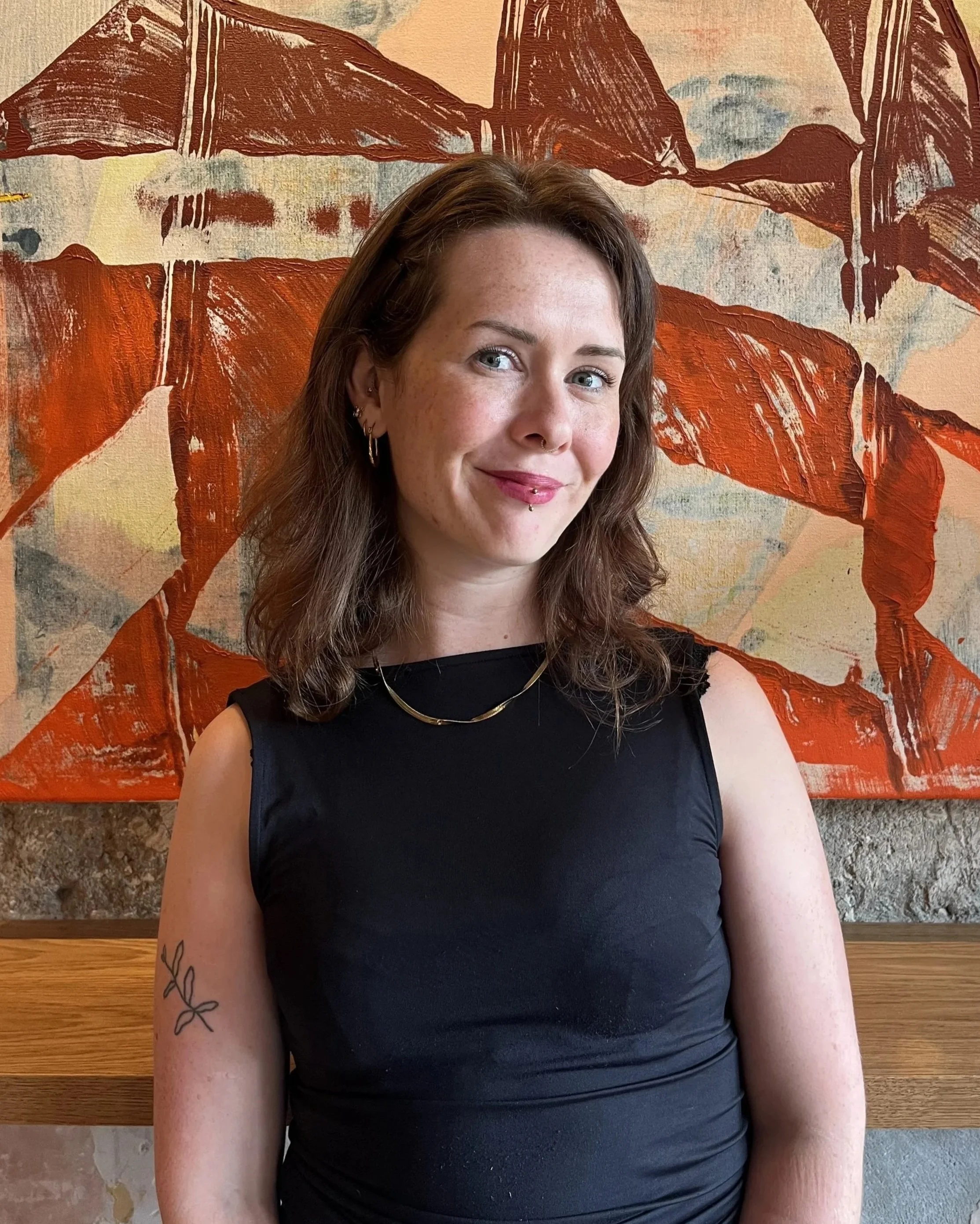 A young woman with brown hair and piercings smiling, wearing a black sleeveless top and gold jewelry, standing against a textured abstract painting with orange and brown tones in a rustic setting.