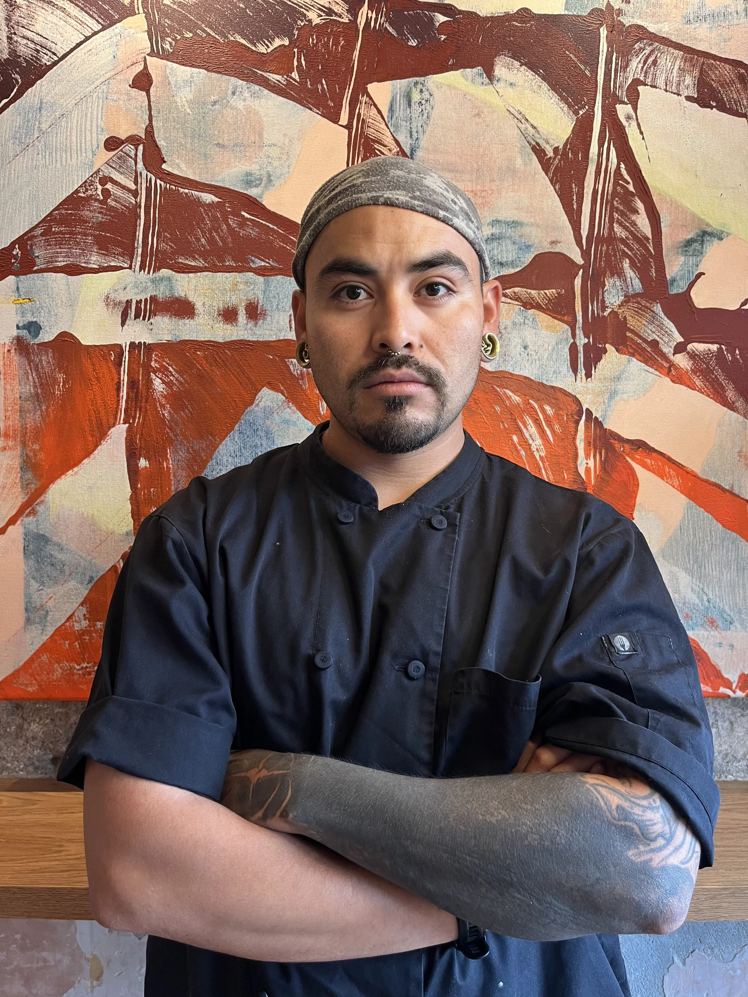 A man with a beard and earrings wearing a grey head covering and black chef's coat, standing in front of an abstract painting with large orange, red, blue, and white brushstrokes.