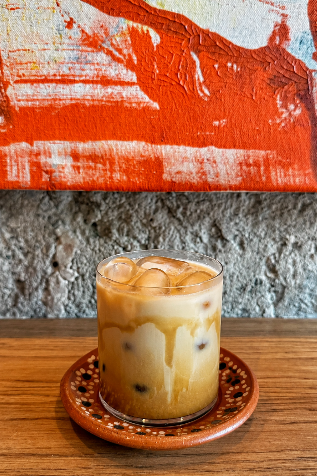 A glass of iced coffee with milk and ice cubes on a wooden table, with a textured stone wall and abstract orange and red artwork in the background.