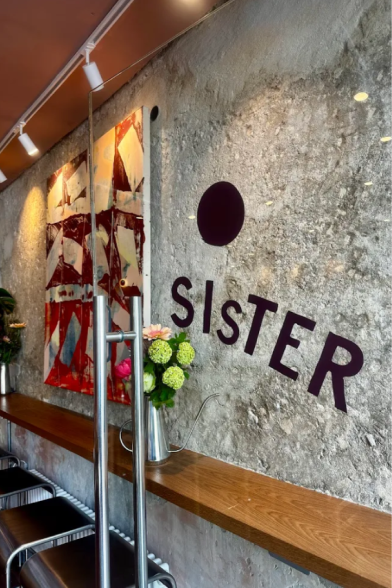 Interior of a room with a stone wall decorated with the word 'SISTER' in black letters, a black circular shape above the text, a colorful abstract painting, a wooden shelf with a flower arrangement, a ladder, and black chairs.