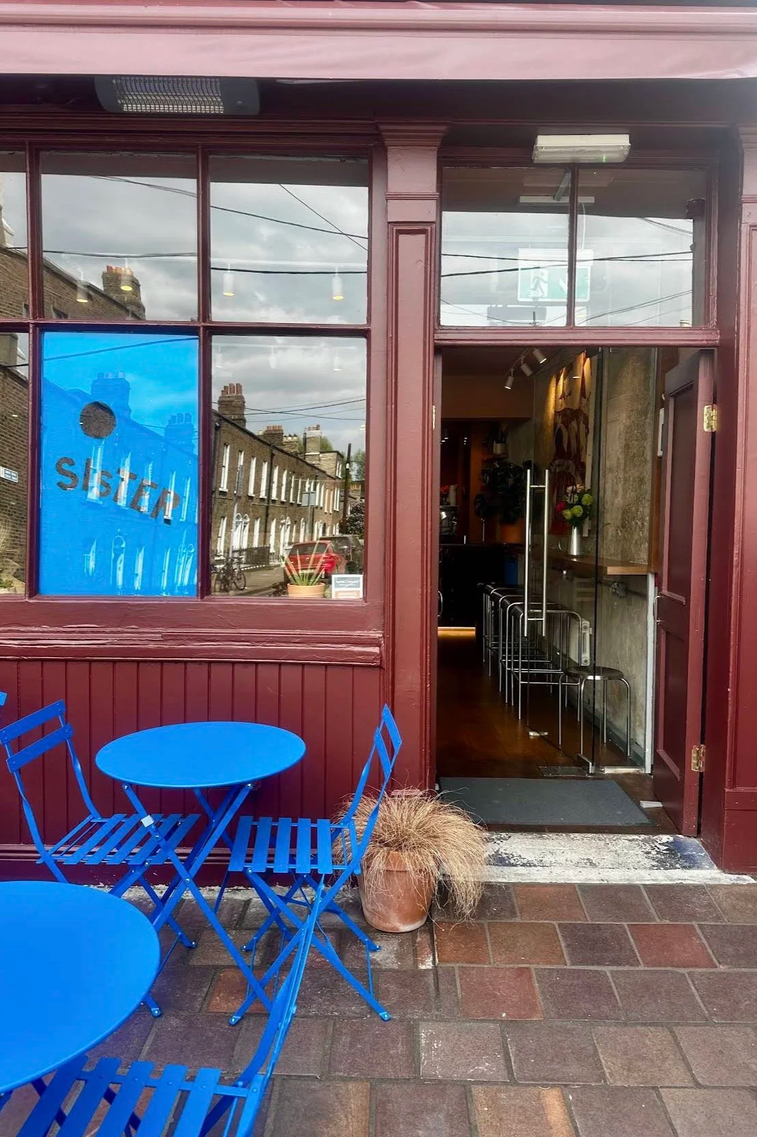 Outside view of a cafe with a maroon exterior, blue outdoor chairs and tables, and a potted plant. The open door shows a glimpse of the interior with bar stools and a decorative wall.