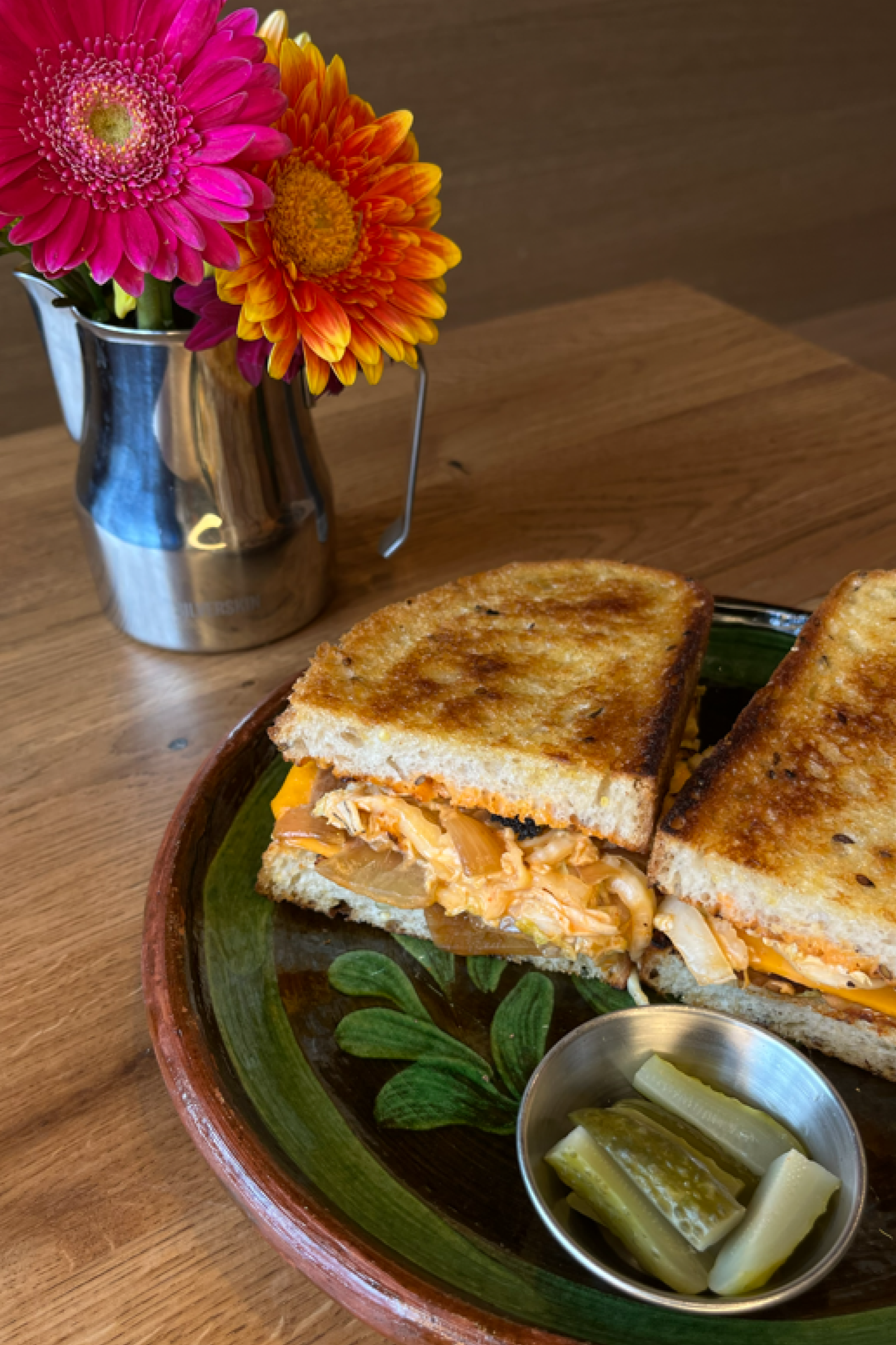 A plate with a grilled chicken sandwich and a side of pickles, served on a rustic ceramic plate with floral patterns. In the background, there is a metal vase with bright pink and orange flowers on a wooden table.