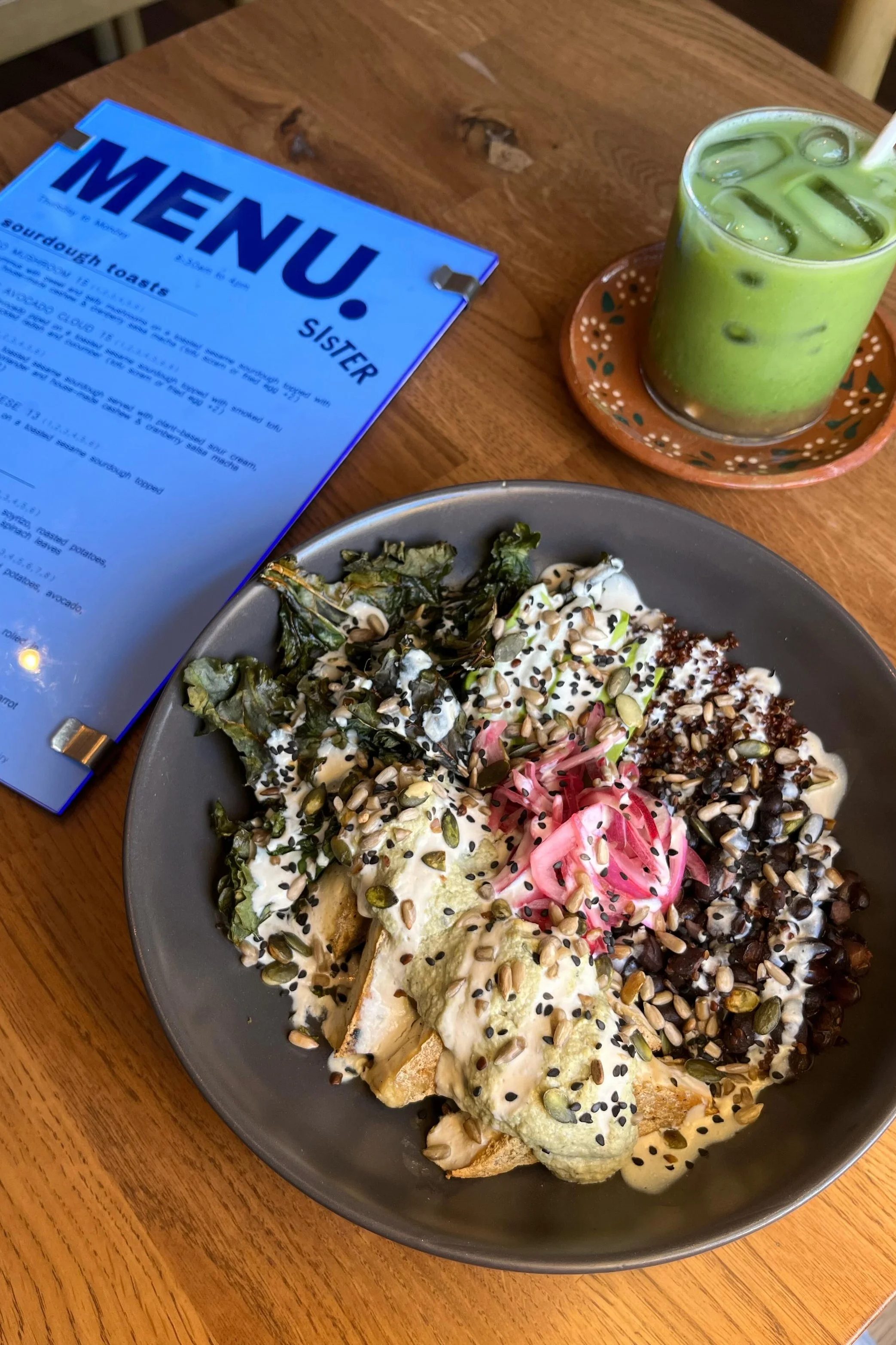 A plate of sliced bread topped with melted cheese, chopped tomatoes, and assorted seeds and herbs, served on a wooden table. In the background, there is a glass of green iced beverage with ice cubes on a small decorative plate, and a menu titled 'MEN