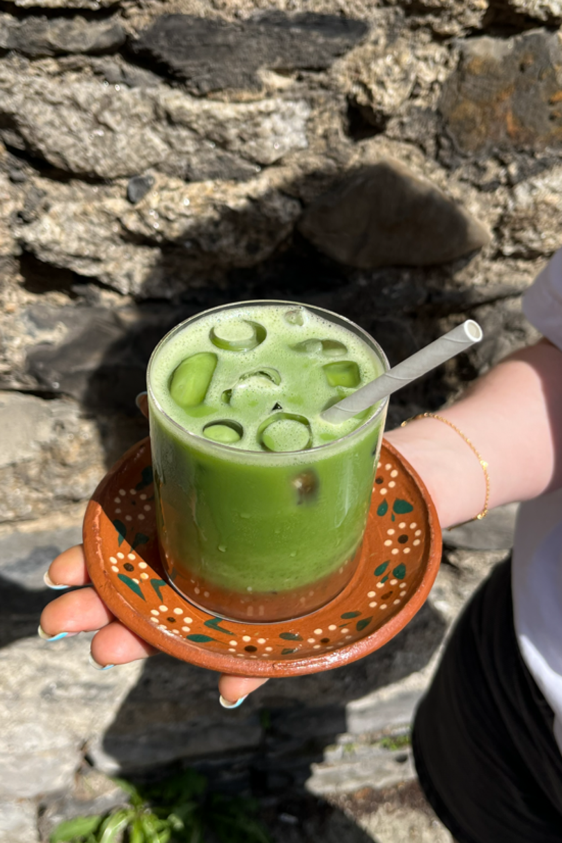 A glass of green iced beverage with ice cubes and lime slices being held on a decorative orange tray in front of a stone wall.