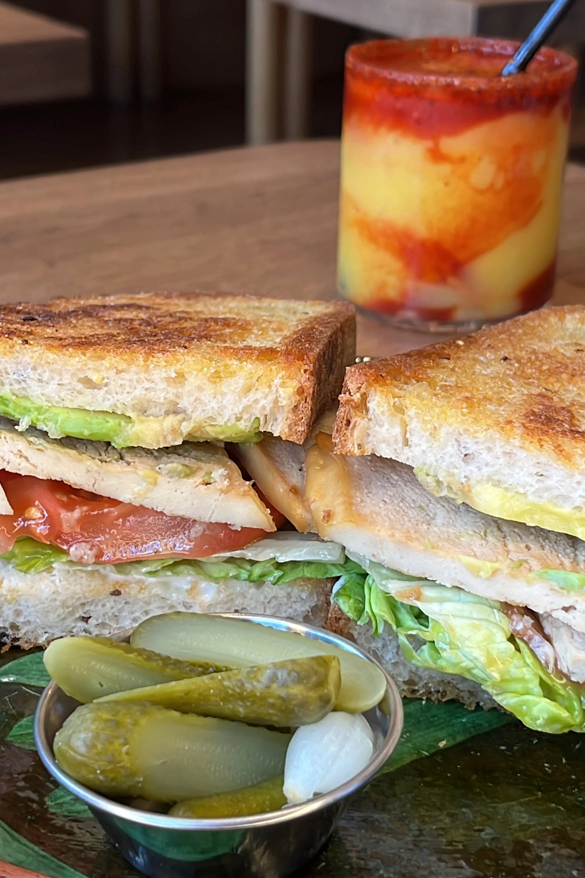 A close-up of a sandwich with toasted bread, lettuce, tomato, and cheese. In the background, a glass jar filled with sliced peaches and syrup, with a spoon inside, and a small bowl of pickles and onions in the foreground.