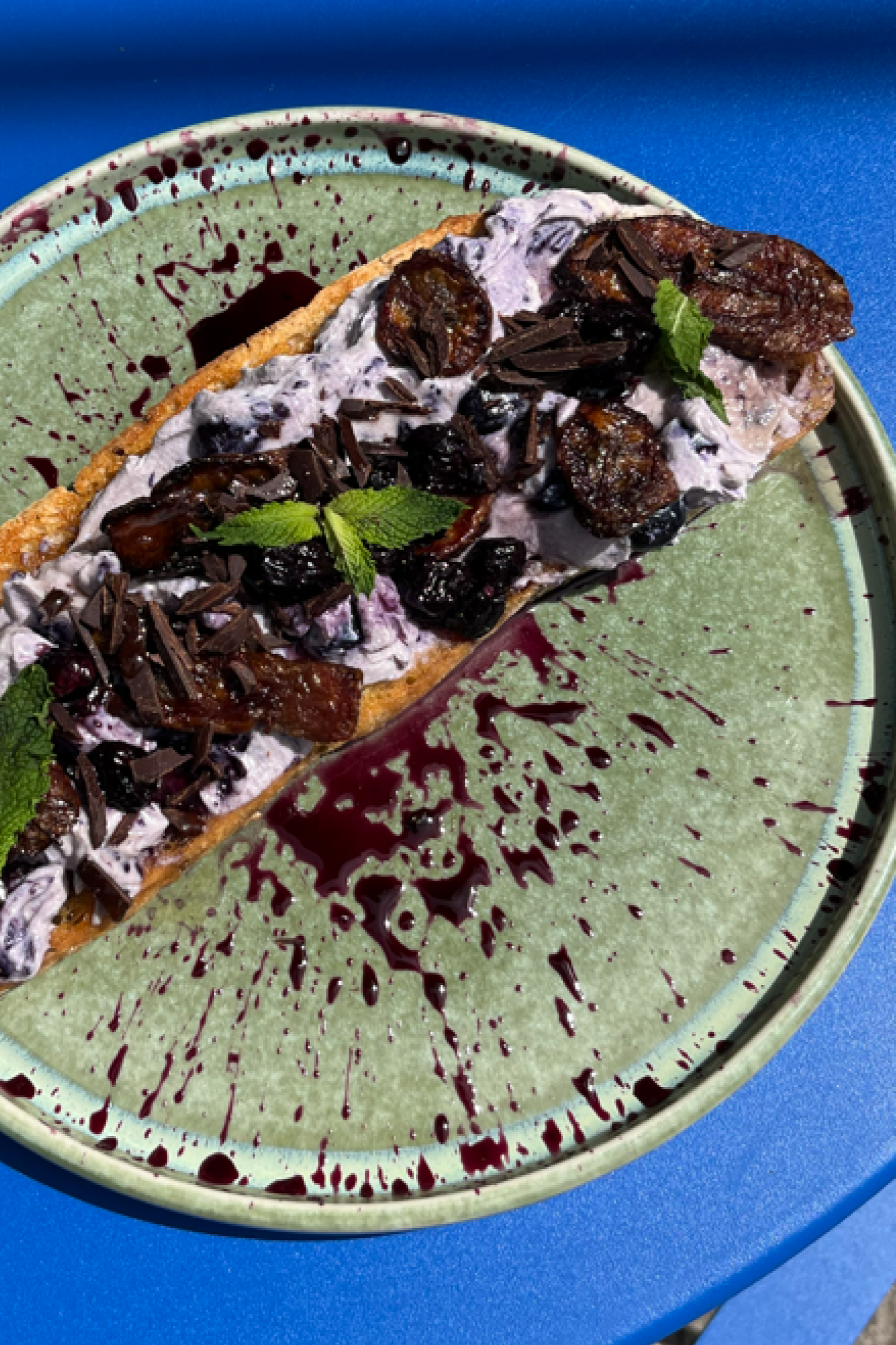 A dessert featuring a chocolate tart with a berry compote, whipped cream, chocolate shavings, and fresh mint leaves on a ceramic platter with green glaze and red splatter design, set on a blue table.
