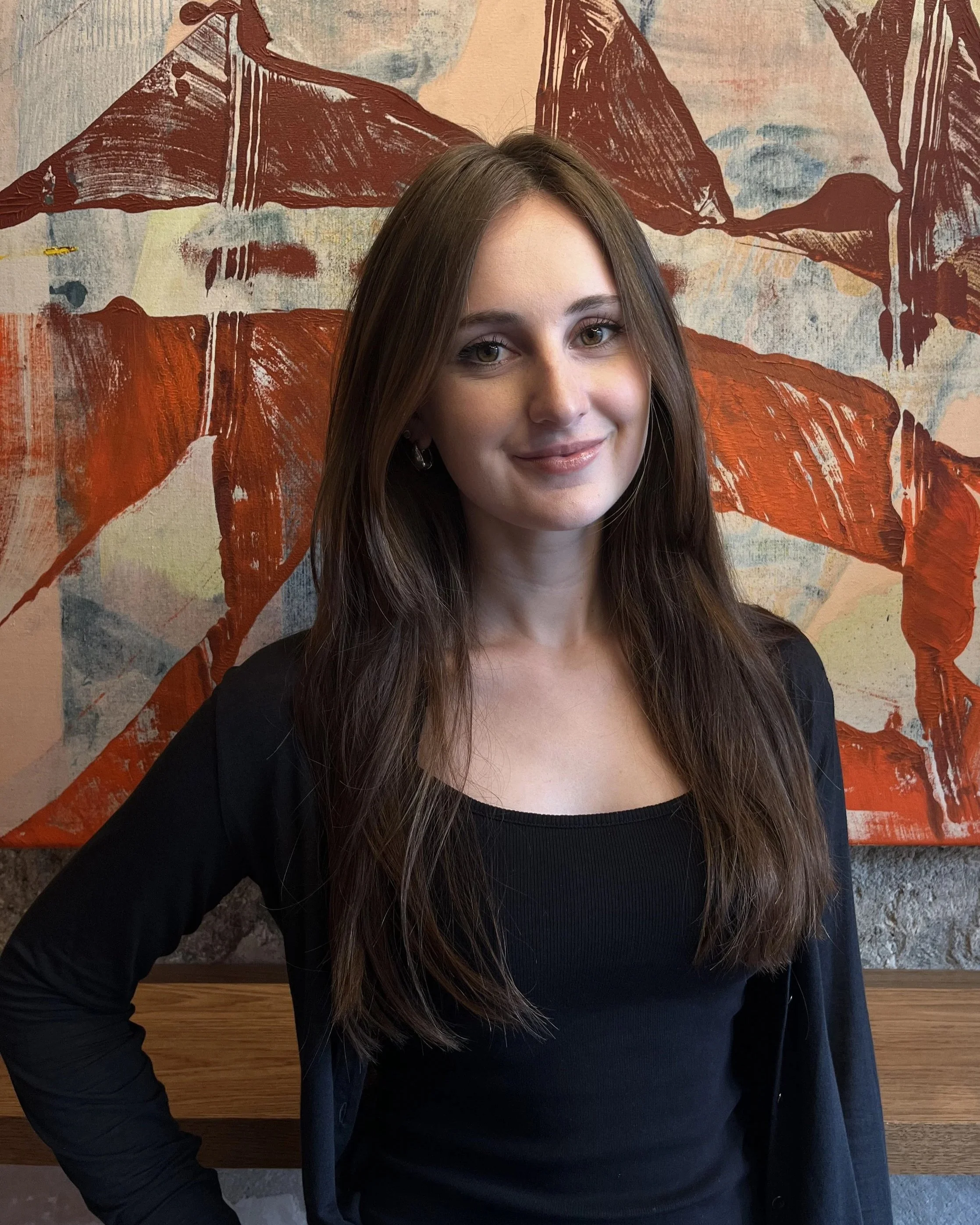 A young woman with long brown hair smiling, wearing a black top and blazer, standing in front of a colorful abstract mural with red, beige, and blue abstract shapes and textures.