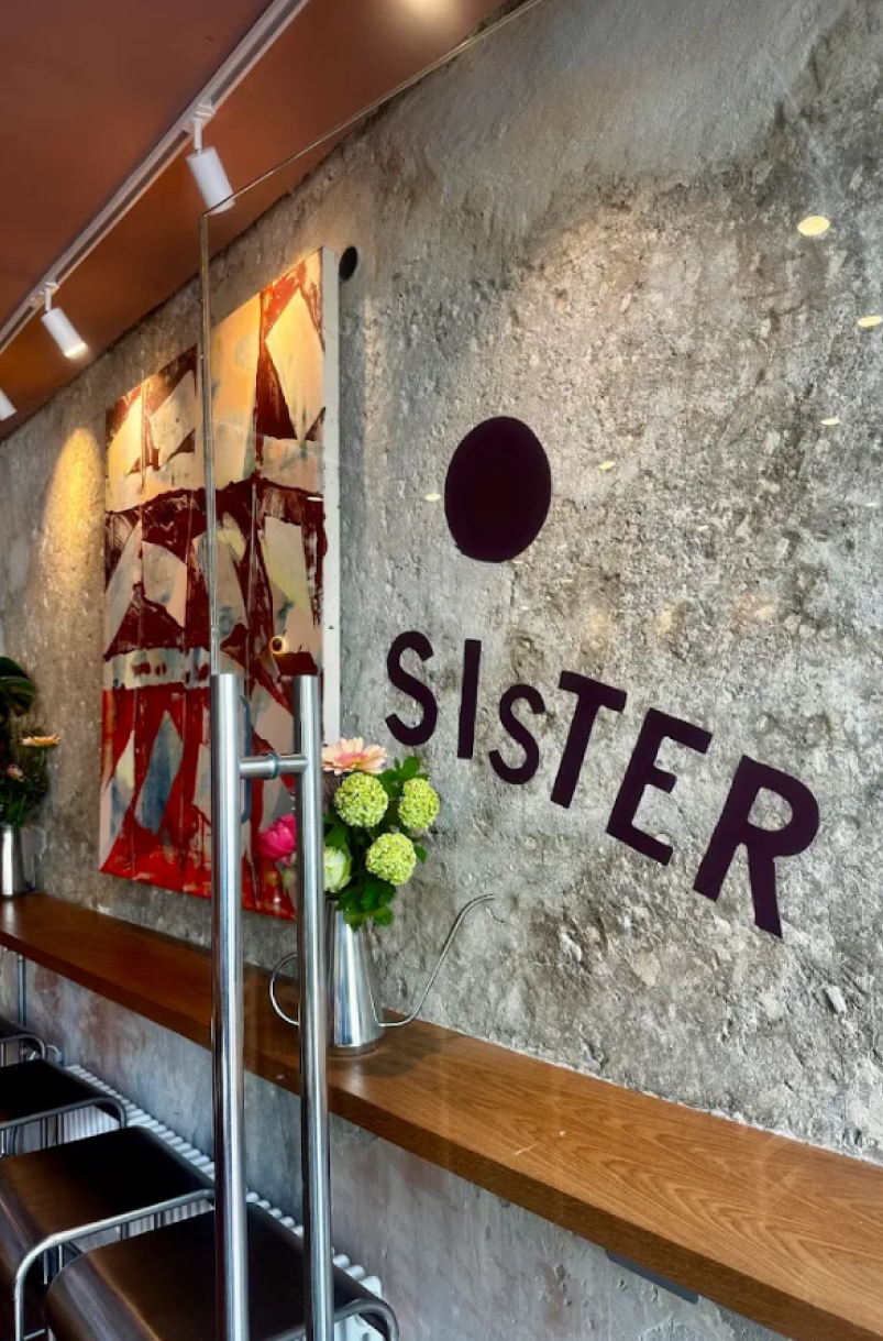 Interior of a cafe or restaurant with a textured stone wall, abstract artwork, a wooden shelf, beige and green flowers in a metallic watering can, black chairs with metal frames, and a decorative sign on the wall that reads 'SISTER' with a black oval