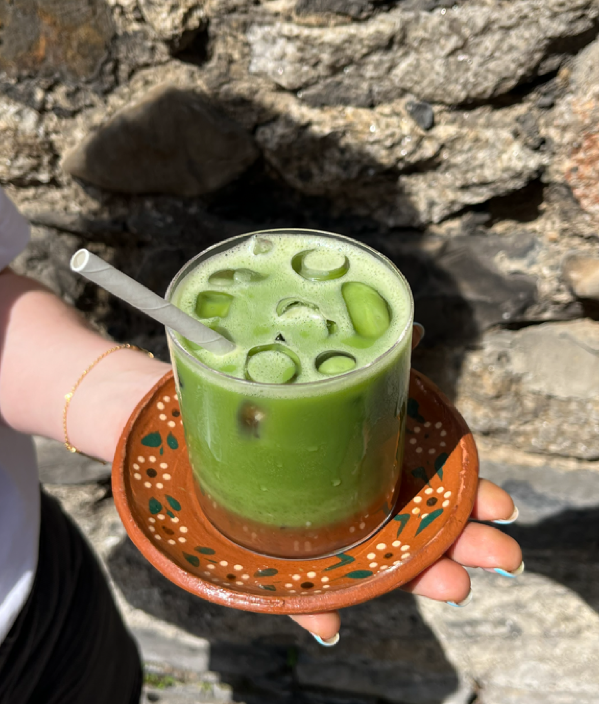 A hand holding a glass of green beverage with ice cubes and lime slices, placed on a decorative terracotta tray with painted designs, against a backdrop of rocky stone surface.