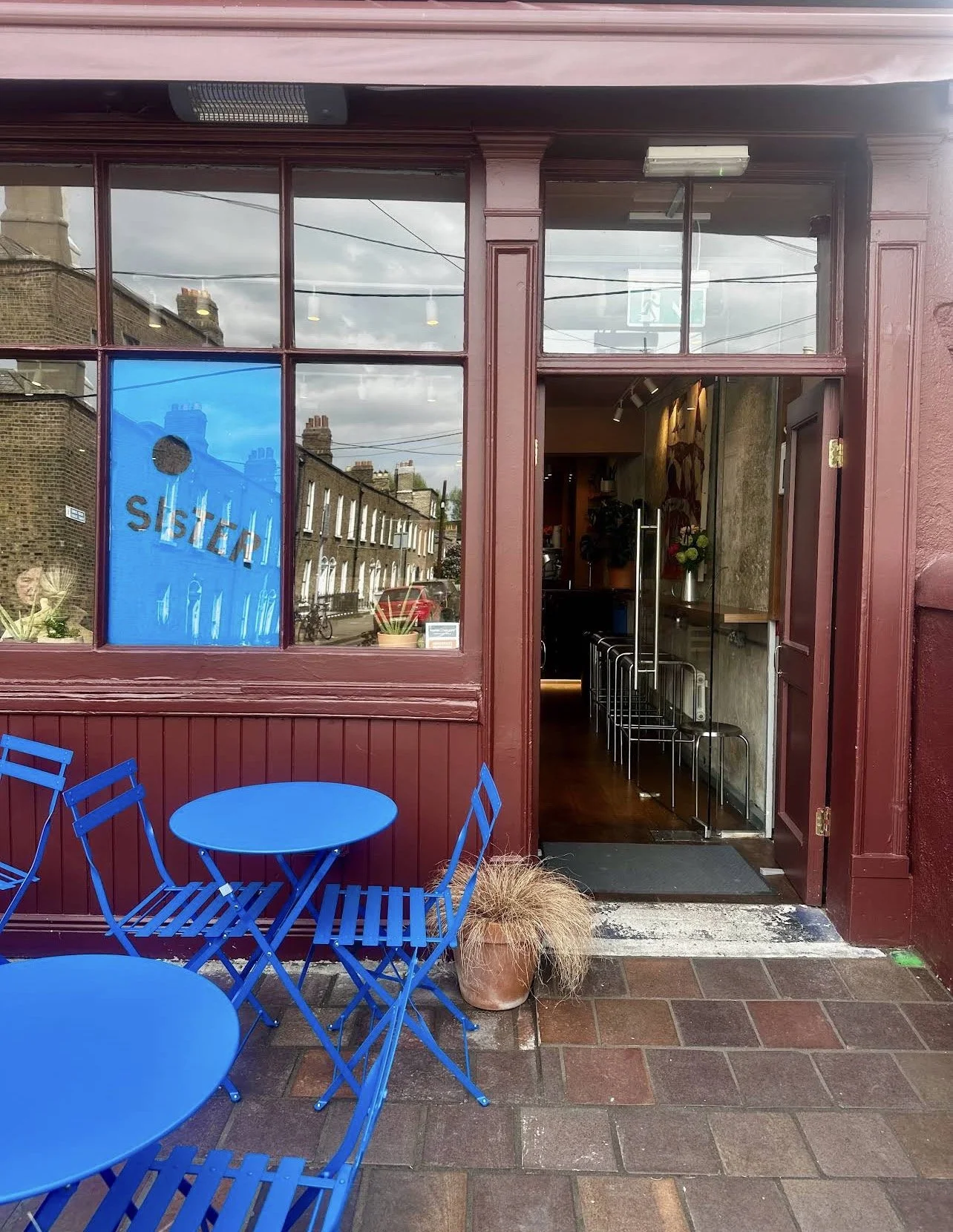 Outside of a cafe with a maroon facade, blue metal tables and chairs on the sidewalk, and a potted plant with brown foliage. The cafe entrance has a glass door and large windows with reflections of buildings and the street. Inside, there are bar stoo
