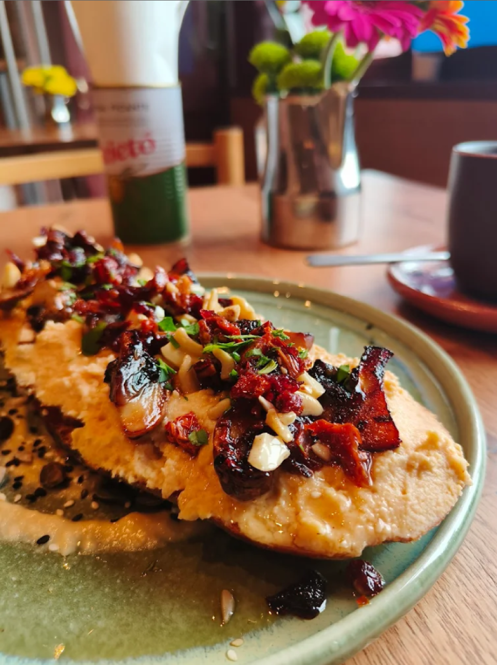 Close-up of a plate of food with a grilled chicken breast topped with chopped bacon, herbs, and sun-dried tomatoes on a green ceramic plate. In the background, a metal jar with pink and orange flowers, a dark mug on a saucer, and a can of food are visible on a wooden table.