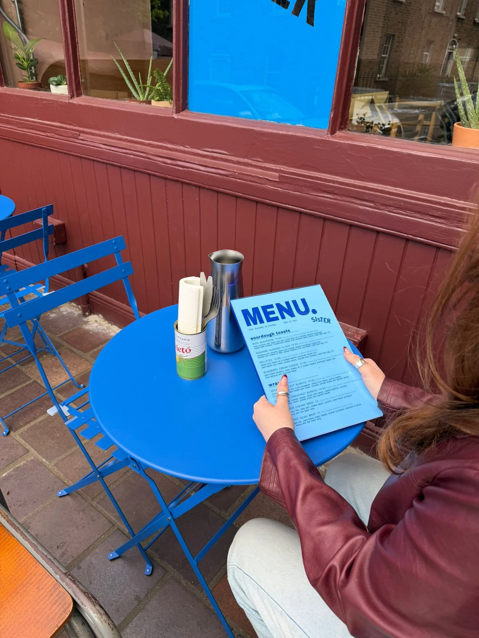 A person holding a blue menu card at an outdoor seating area, with a blue round table, a small canister with rolled paper napkins, and blue chairs. There are potted plants behind a red wooden wall and windows reflecting a street scene.