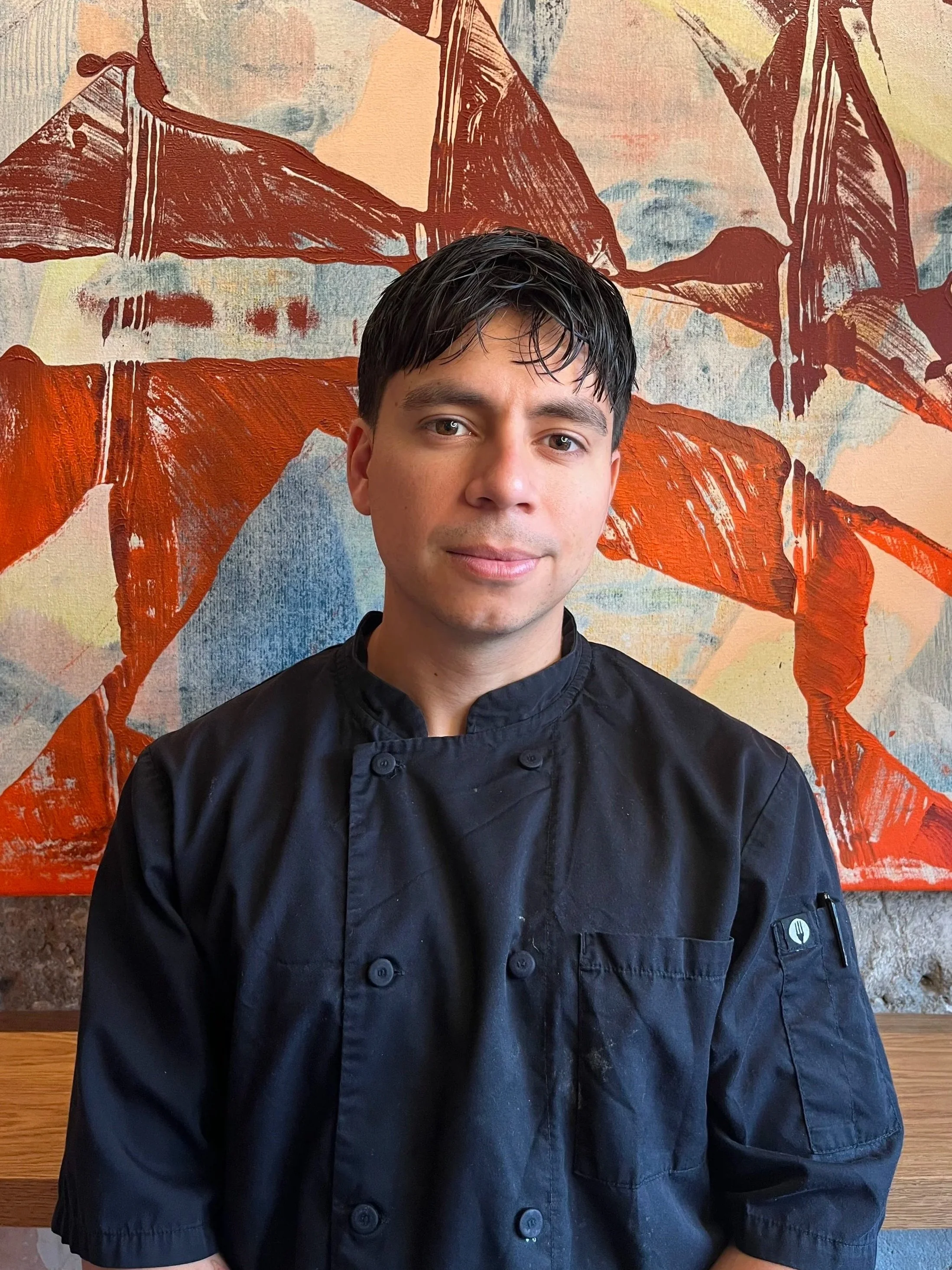 A young man with short black hair and fair skin, wearing a black chef's jacket, standing in front of an abstract painting featuring large orange and brown shapes on a textured multicolored background.